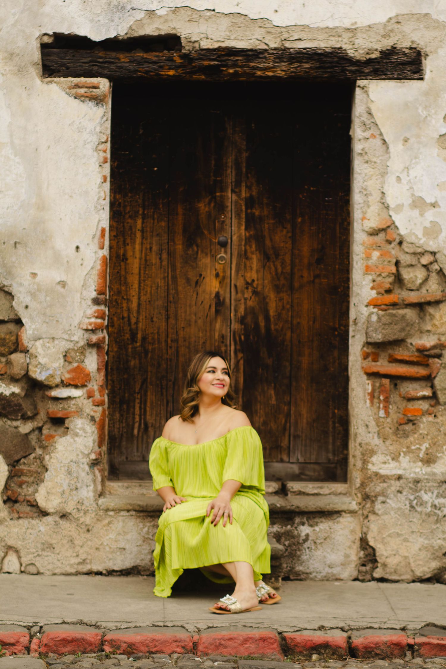 A bright lime green dress contrasts against a rustic wooden doorway and weathered stone wall.