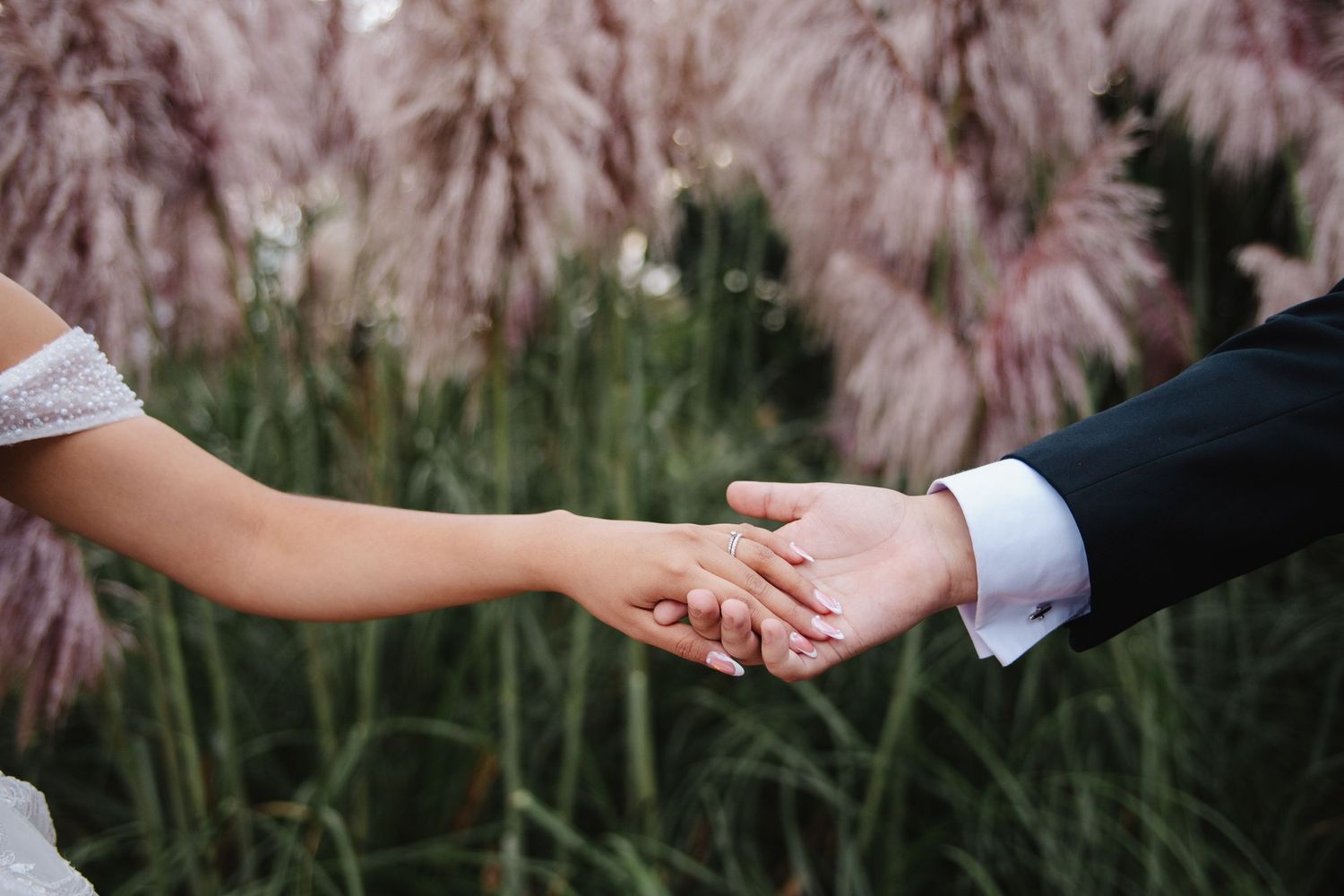 Bride and groom holding hands outdoors with soft pink pampas grass in the background at a wedding.