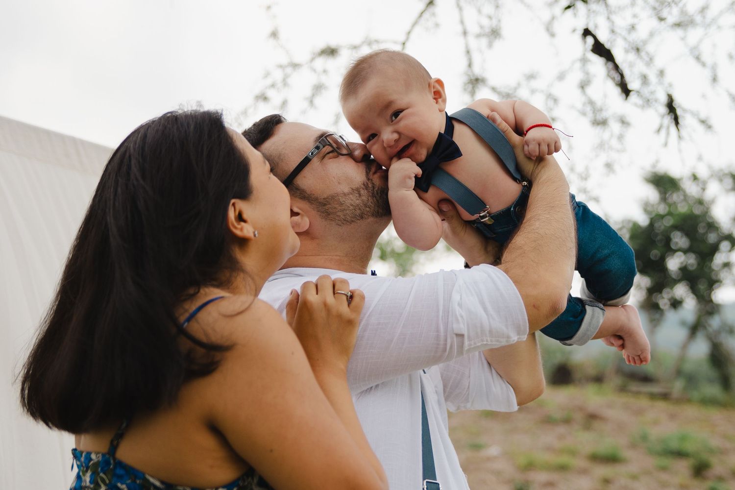 Happy parents kissing their smiling baby outdoors, sharing a joyful family moment together.