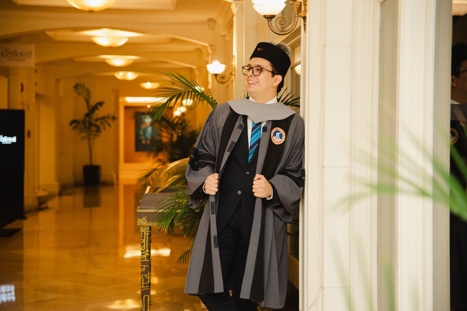 Graduate in academic gown and mortarboard poses proudly in elegant hall with marble columns and warm lighting.