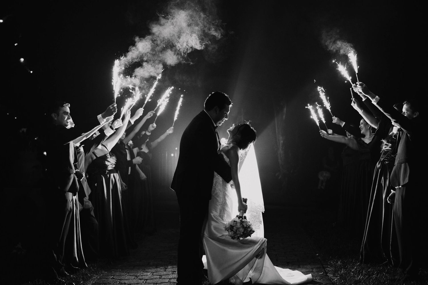 Dramatic black and white night wedding photo with sparklers forming tunnel of light.