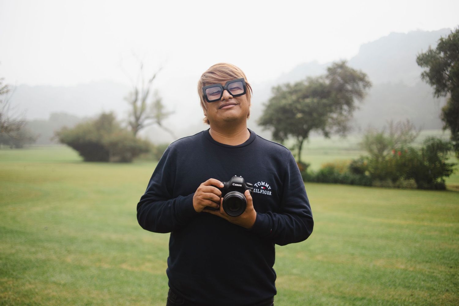 A person in sunglasses and dark clothing holds a camera while standing in a misty green field.