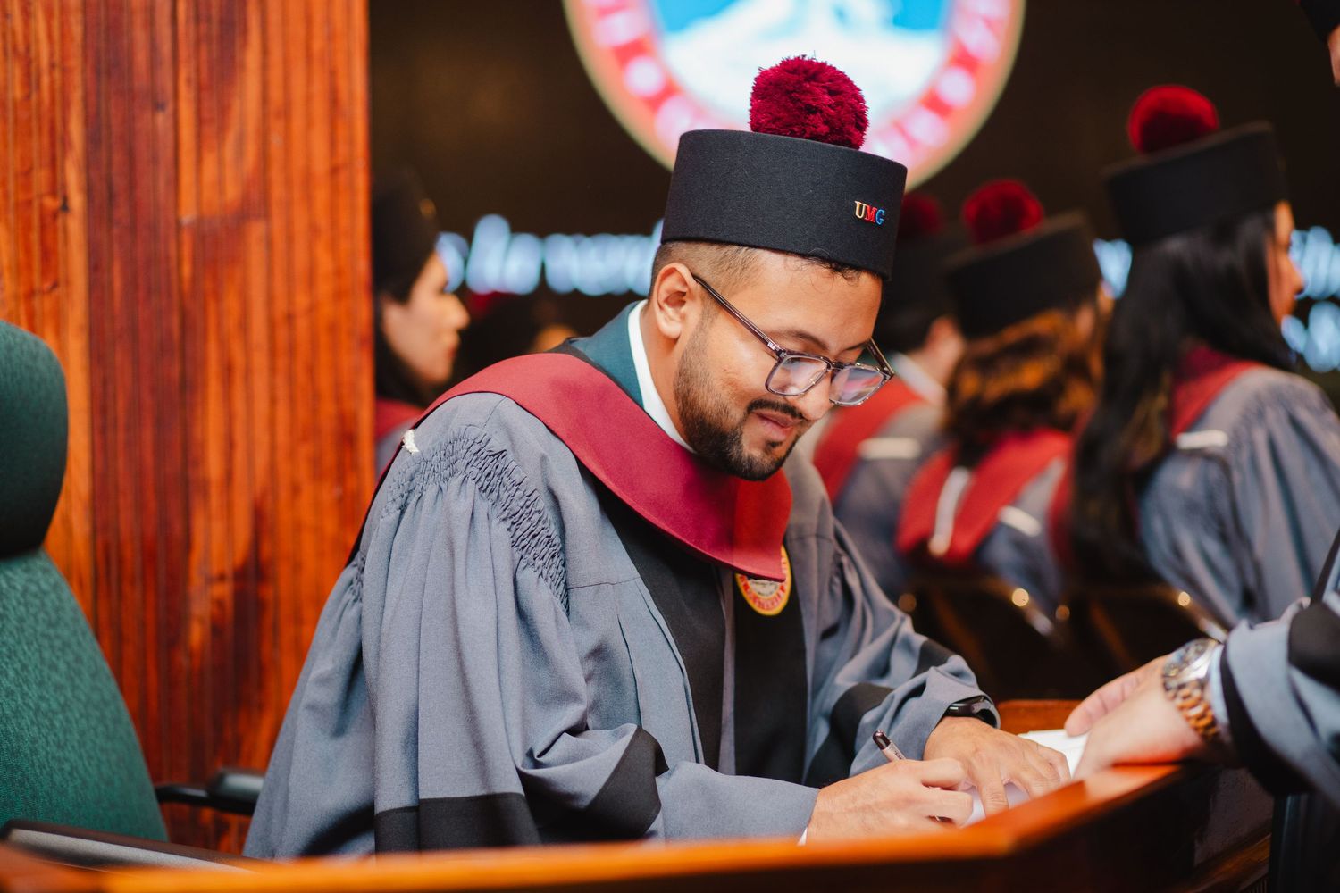 A row of graduates in academic regalia sit during a graduation ceremony in a wood-paneled auditorium.