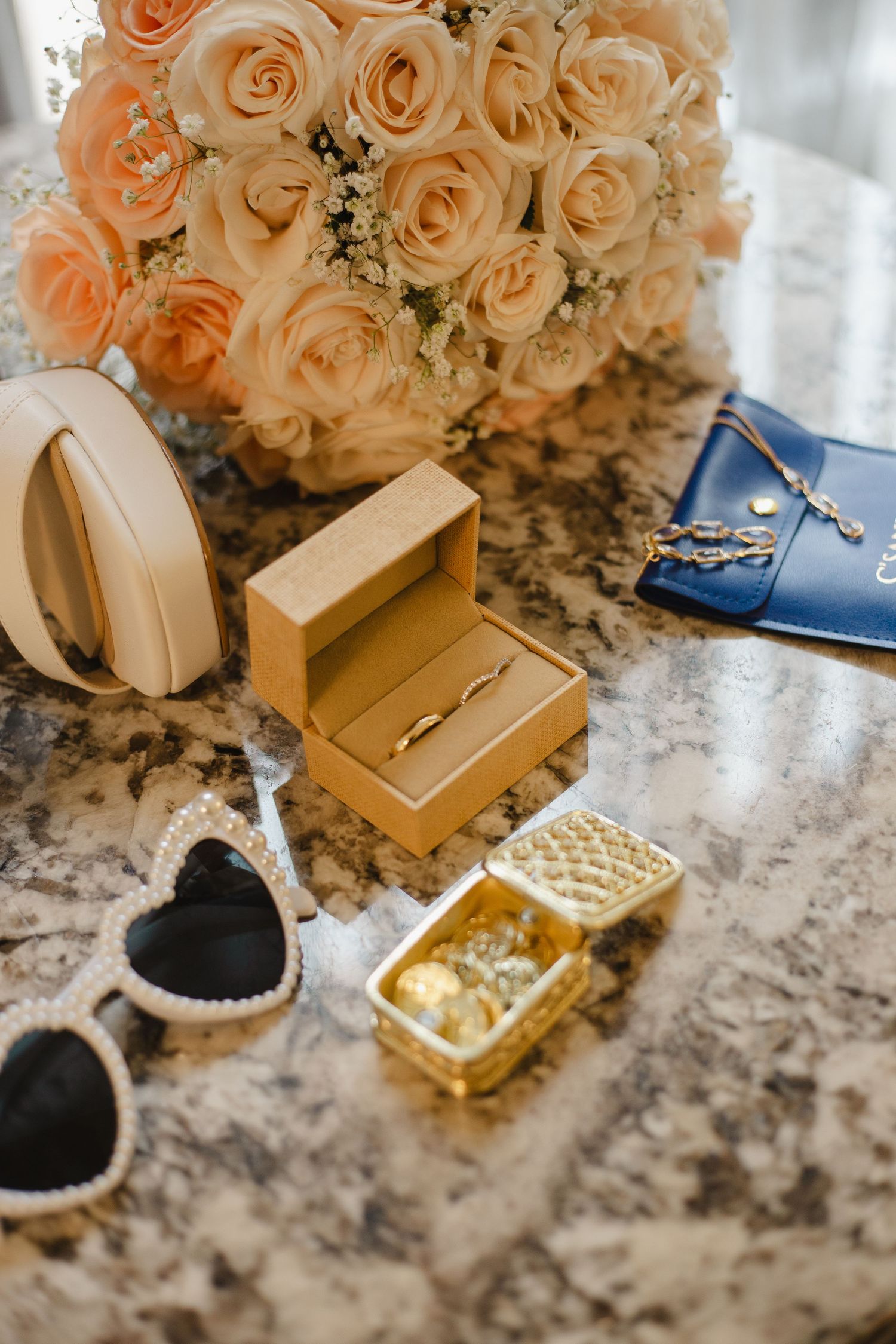 Wedding accessories including peach roses, jewelry boxes, and blue shoes arranged on a marble surface.