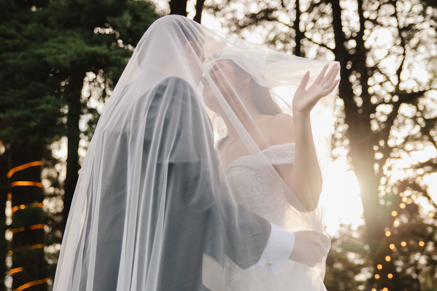 Two brides sharing a kiss under a flowing white veil in a sunlit forest setting on their wedding day.