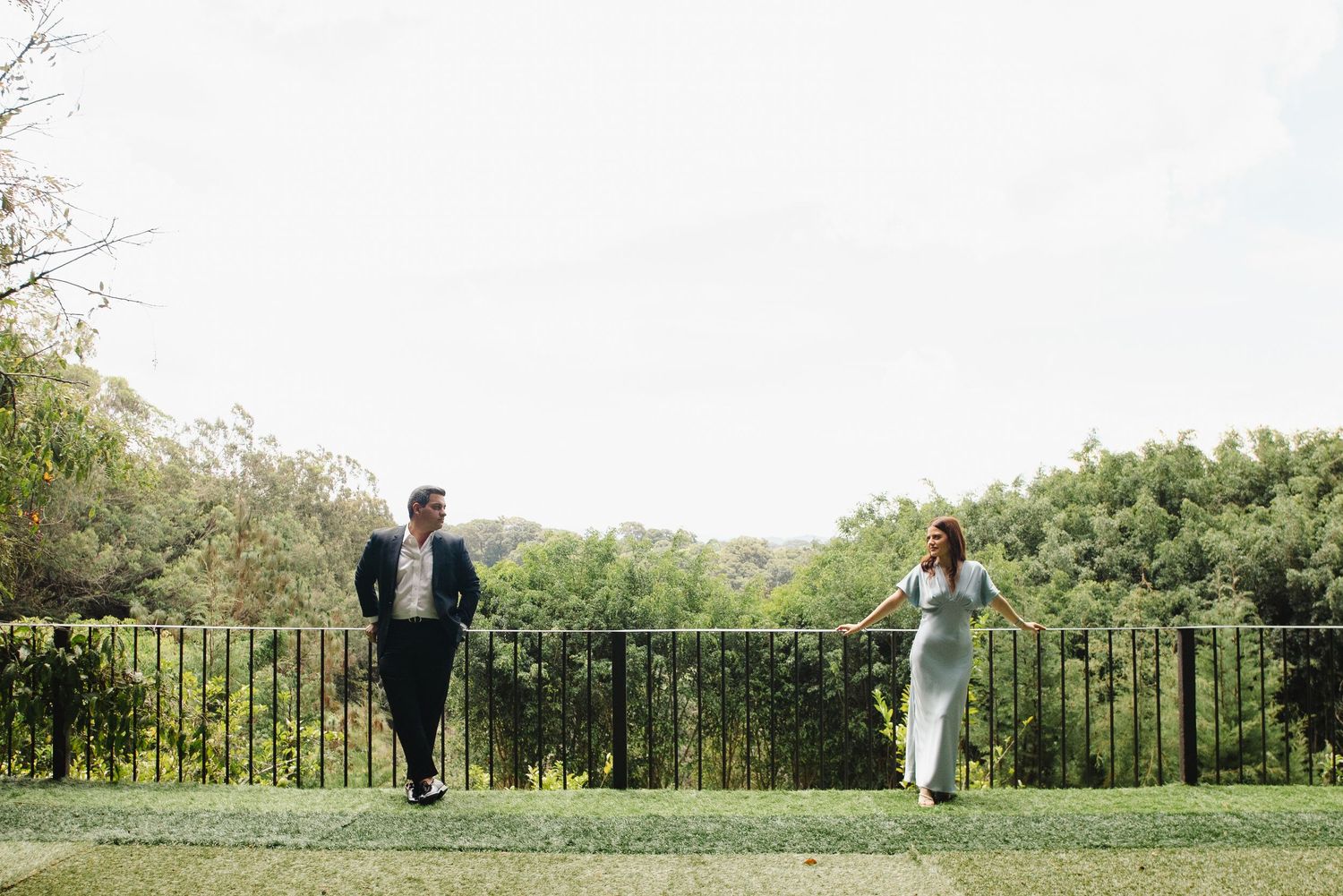 A couple in formal attire walks across a lush green lawn with an iron railing and trees in the background.
