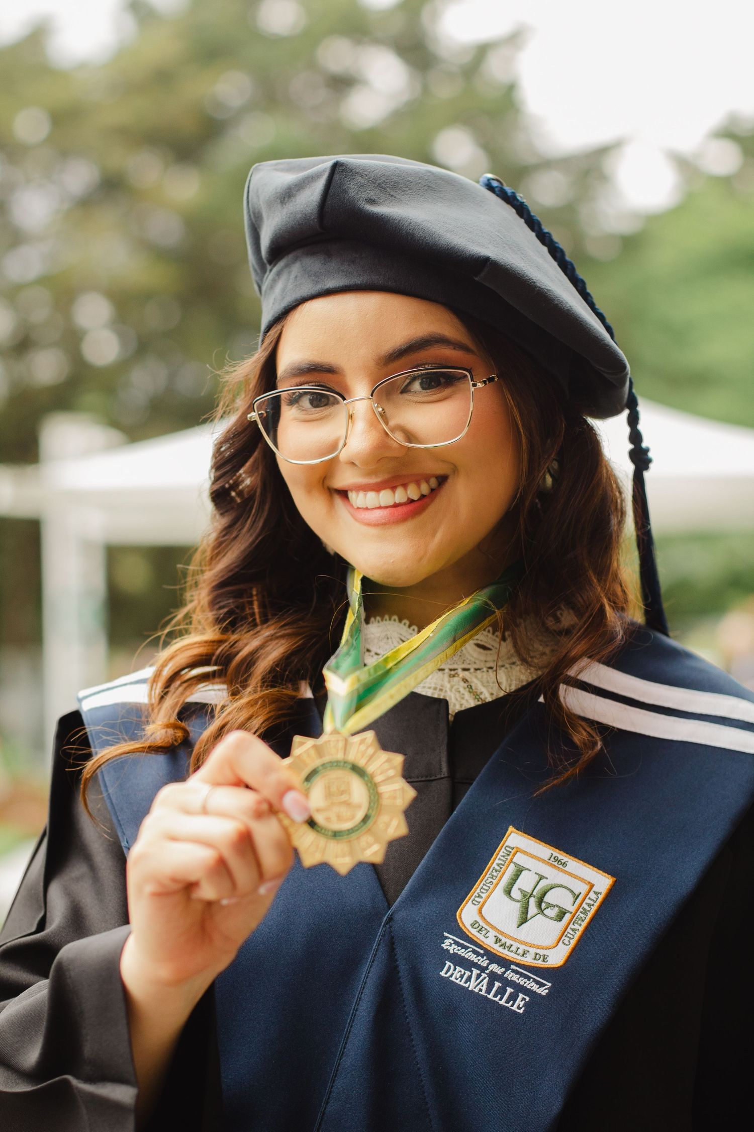 Graduate in cap and gown holding up gold medal achievement award at outdoor graduation ceremony.