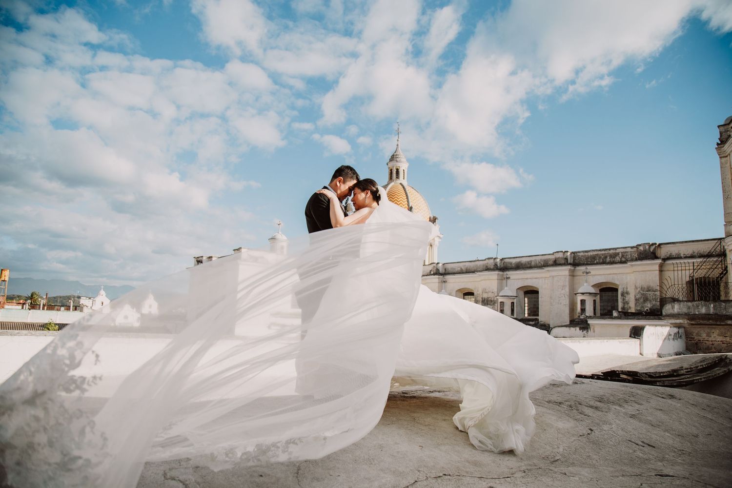 Dramatic wedding photo with flowing veil against blue sky and white architecture.
