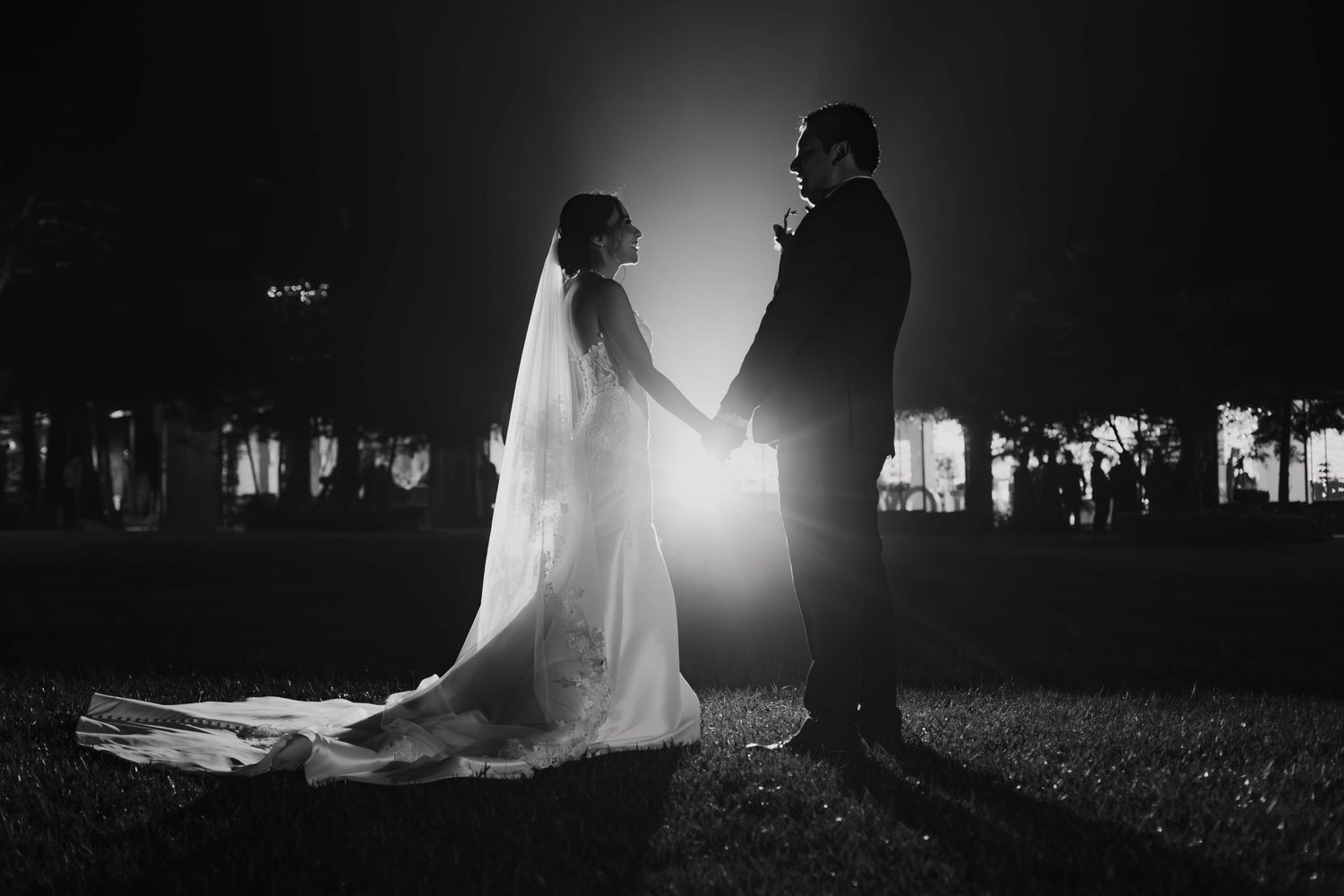 A black and white silhouette of a bride and groom hold hands outdoors at night with a glowing backlight between them.