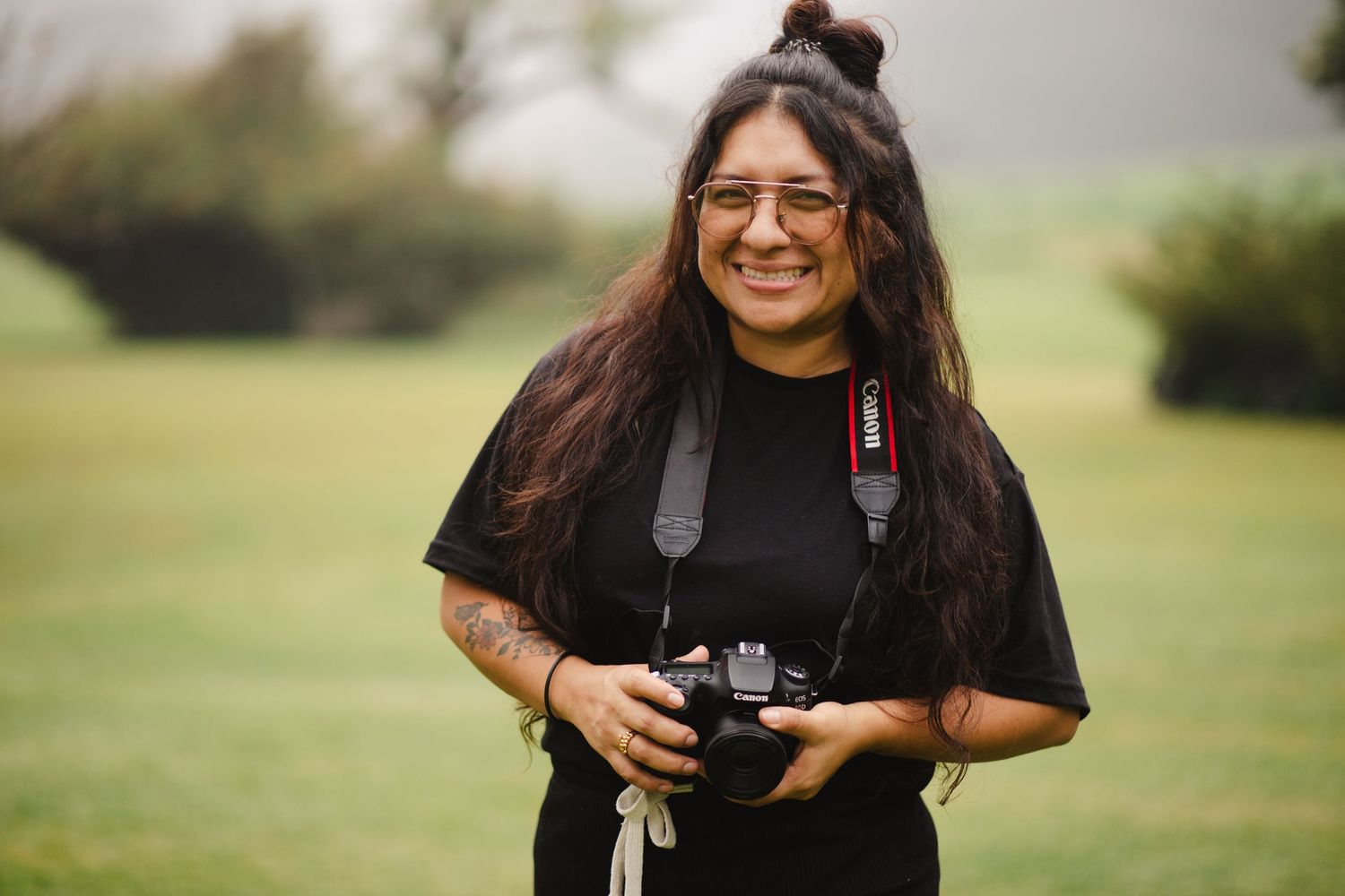 A photographer in a black t-shirt smiles while holding a camera in an outdoor setting with green grass and trees.