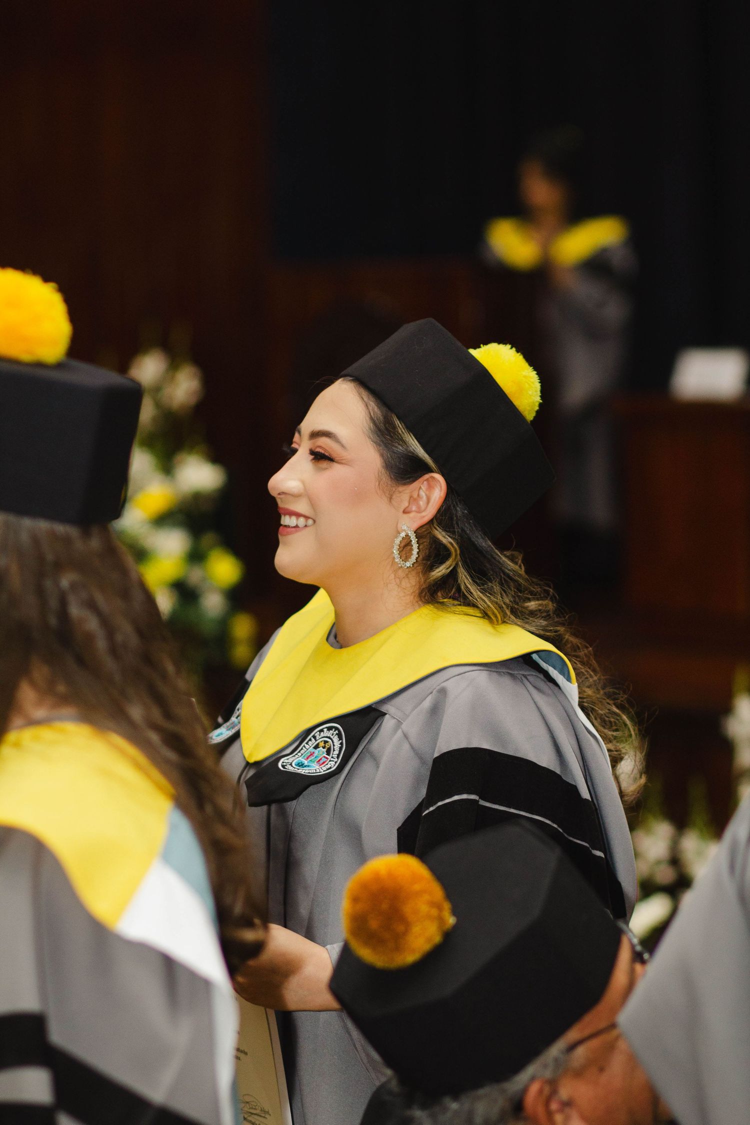 Graduate in grey academic gown with yellow hood smiling during commencement ceremony.