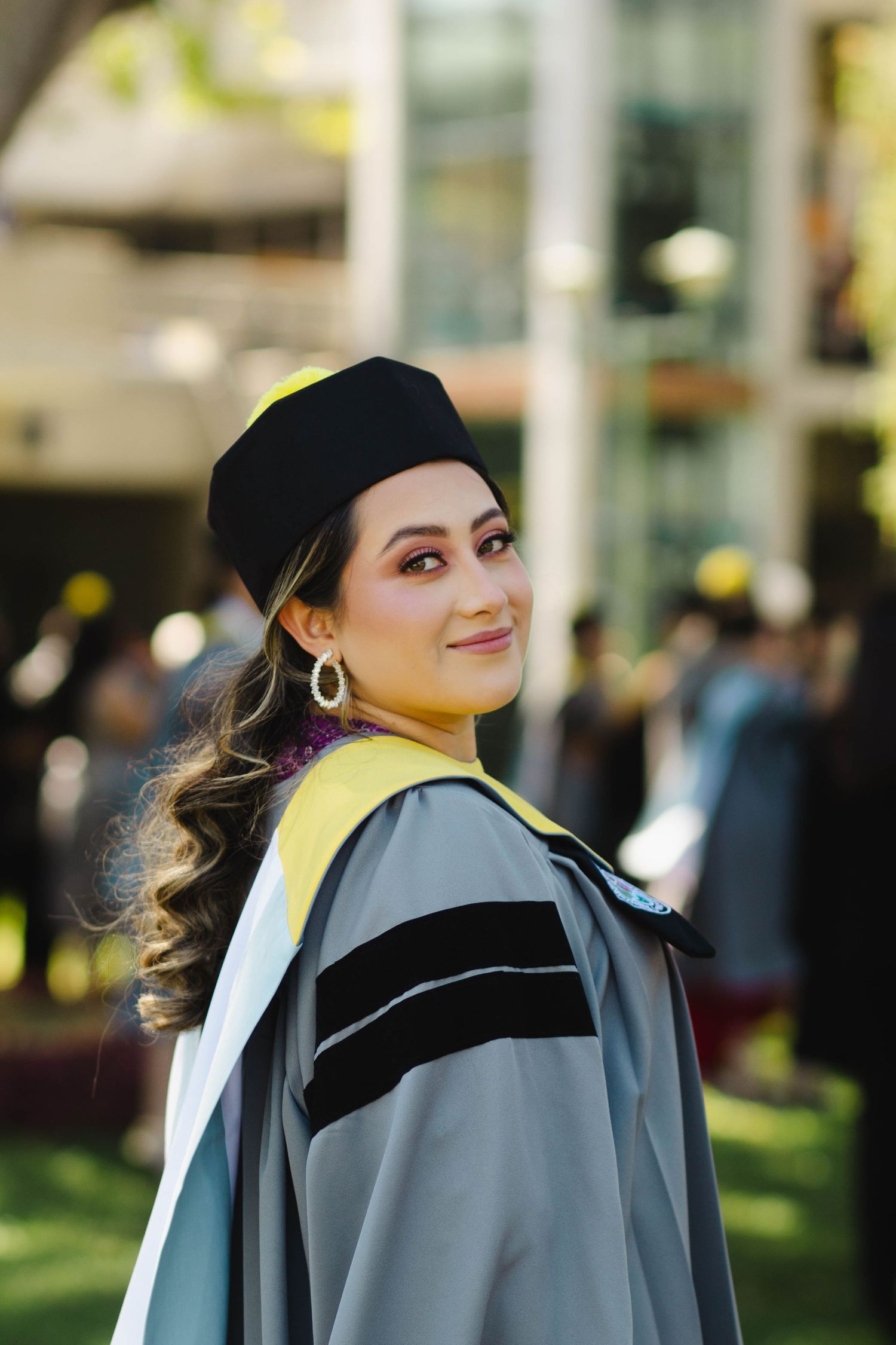 Graduate in academic dress with yellow stole and black cap poses outdoors against blurred background.