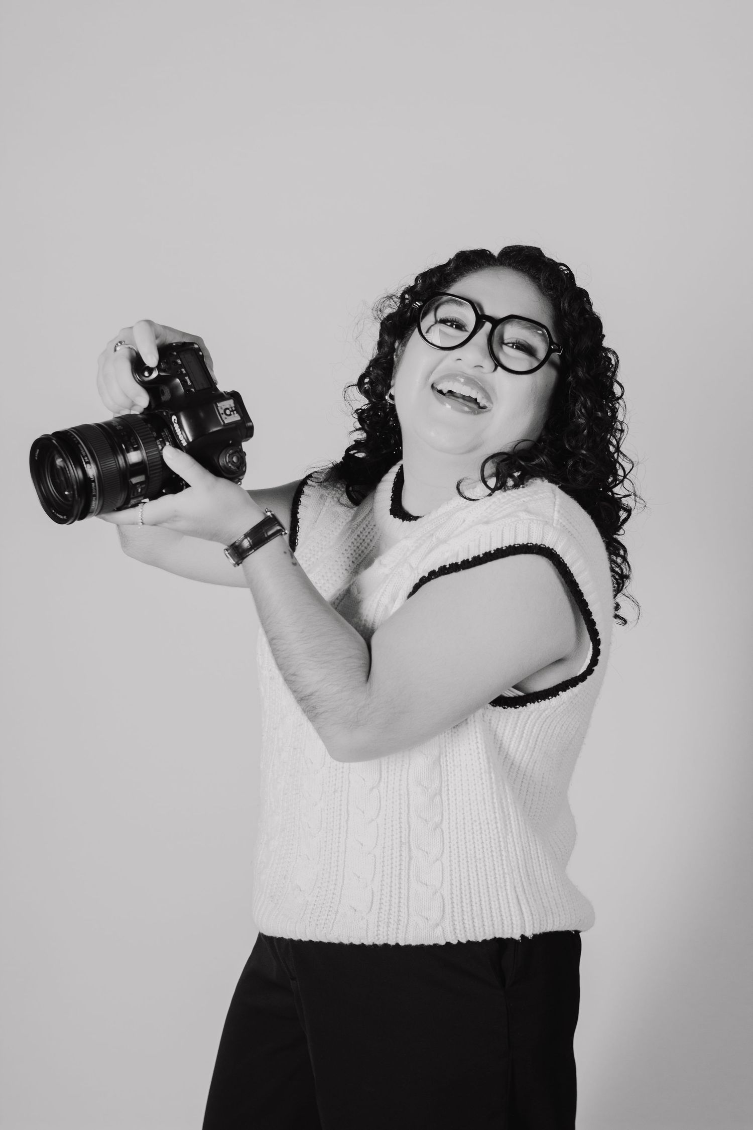 A photographer wearing glasses and a white sweater happily poses with their DSLR camera against a plain background.