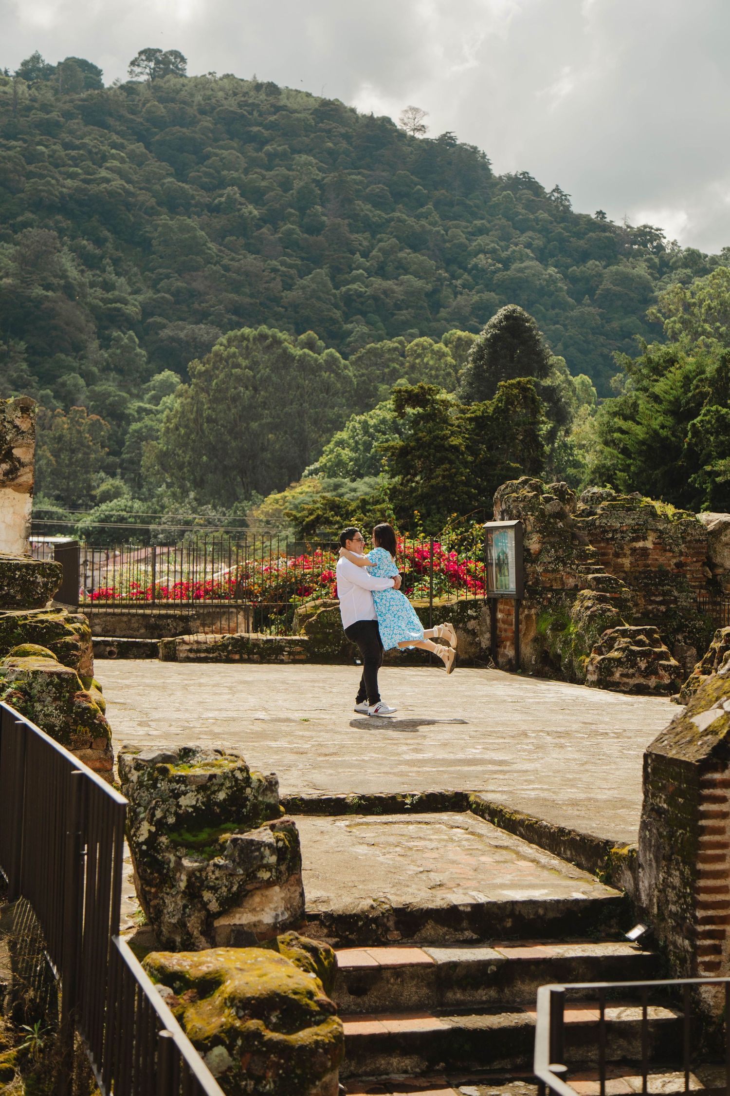 A couple embraces in a romantic pose on stone steps surrounded by ancient ruins and lush green hills.