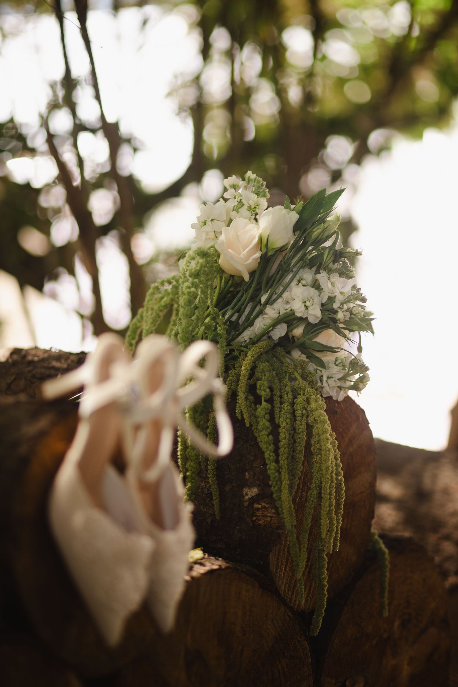 Elegant wedding bouquet of white roses and greenery resting on wooden logs with bridal shoes in a forest setting.