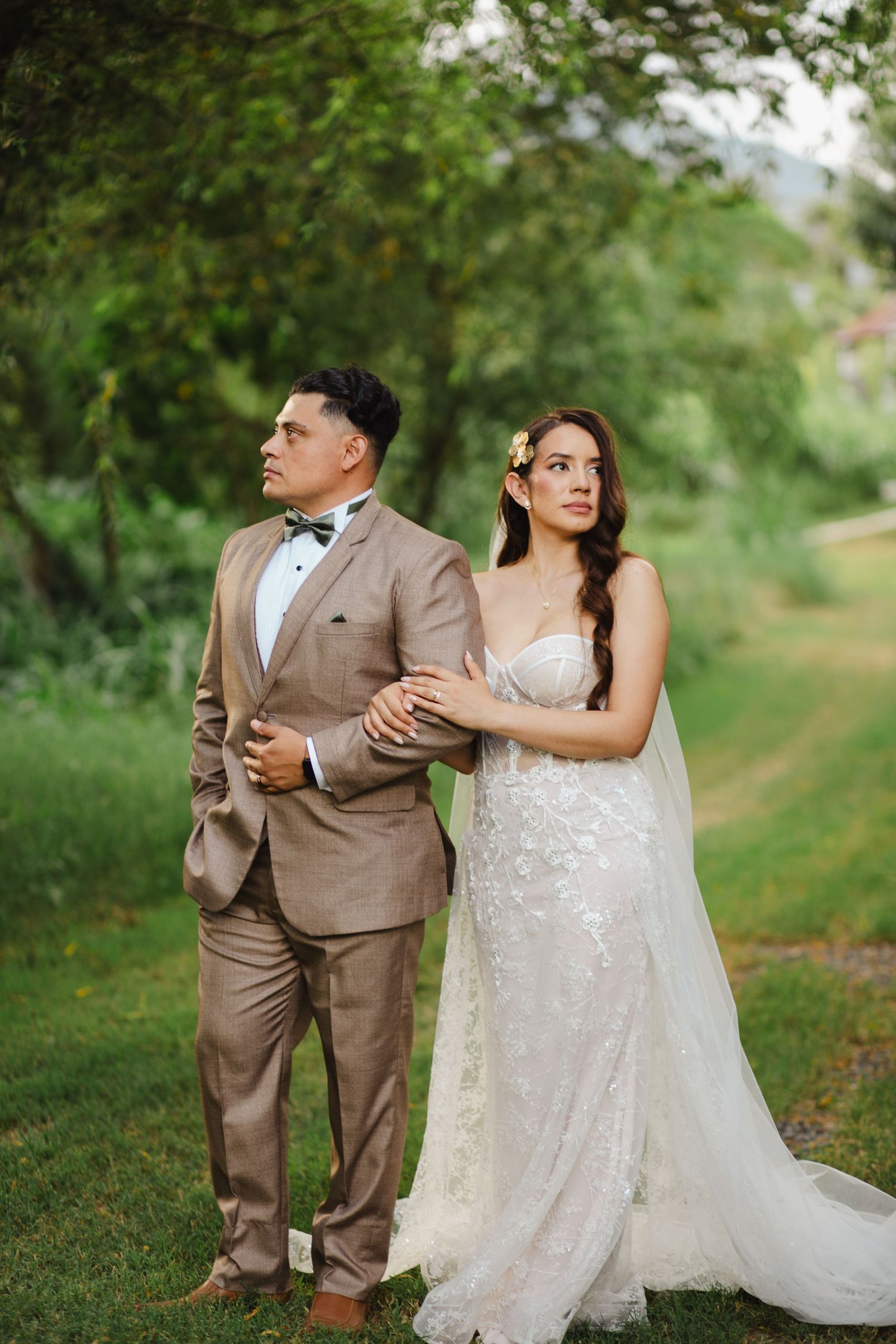 Bride in lace gown and groom in tan suit pose together outdoors on green lawn during elegant garden wedding.