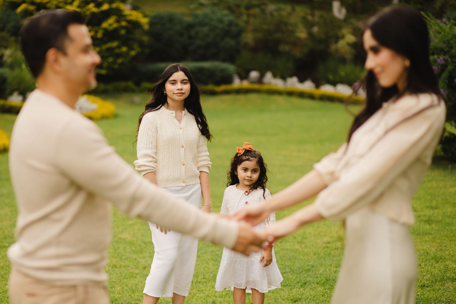 Family of four in matching cream outfits posing outdoors on green lawn, parents holding hands with young daughters.