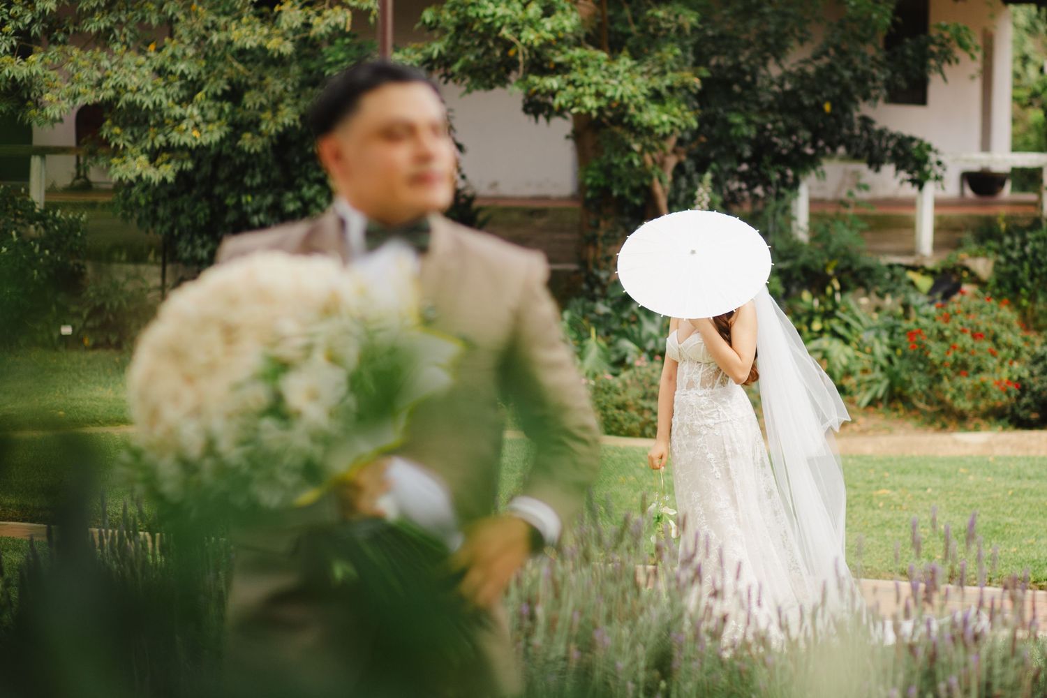 Bride in white gown holding parasol stands in garden as groom holds bouquet in foreground at outdoor wedding.