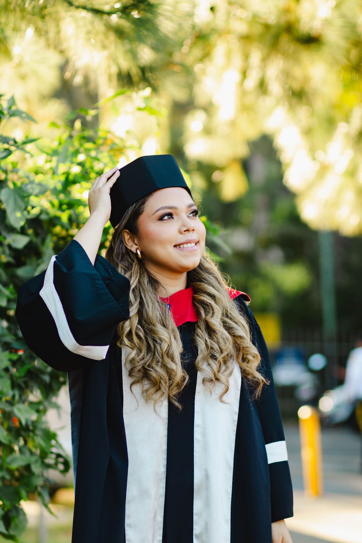 Graduate in black and white academic gown poses outdoors with natural greenery background.