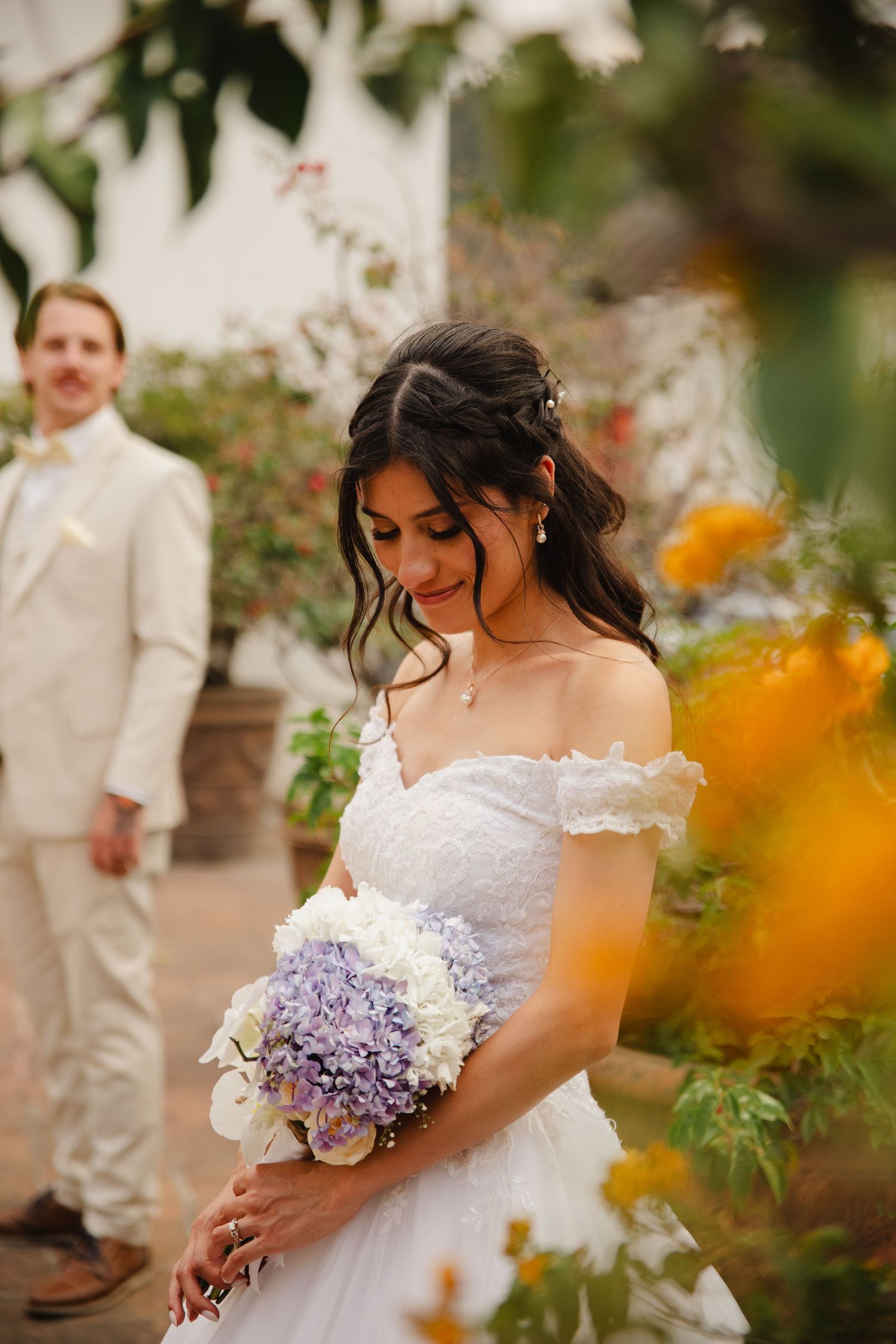 Bride in white off-shoulder gown holds purple hydrangea bouquet, groom in beige suit stands behind in garden.