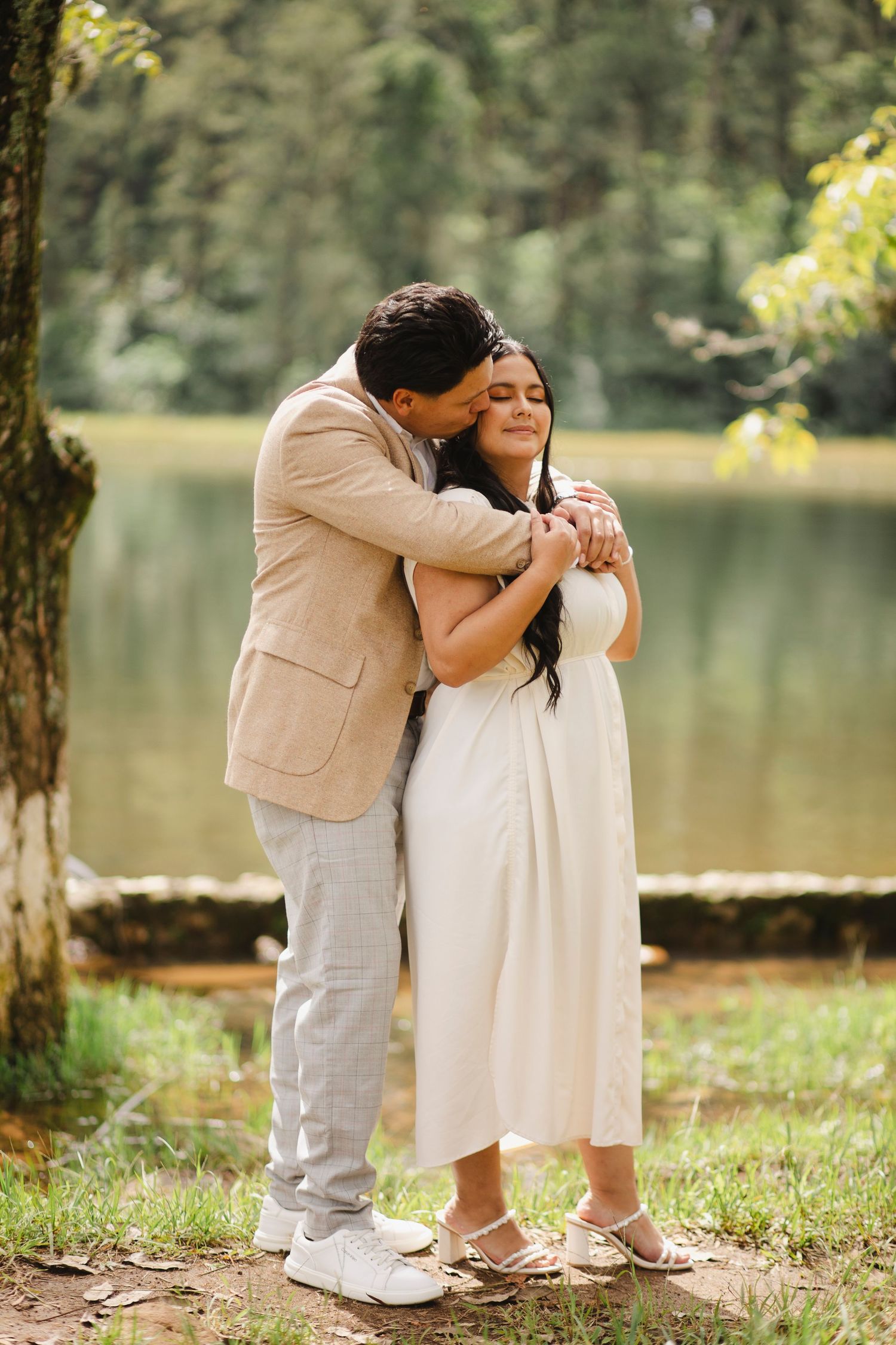 A couple sits together in white and beige outfits by a serene lake surrounded by trees on a sunny day.