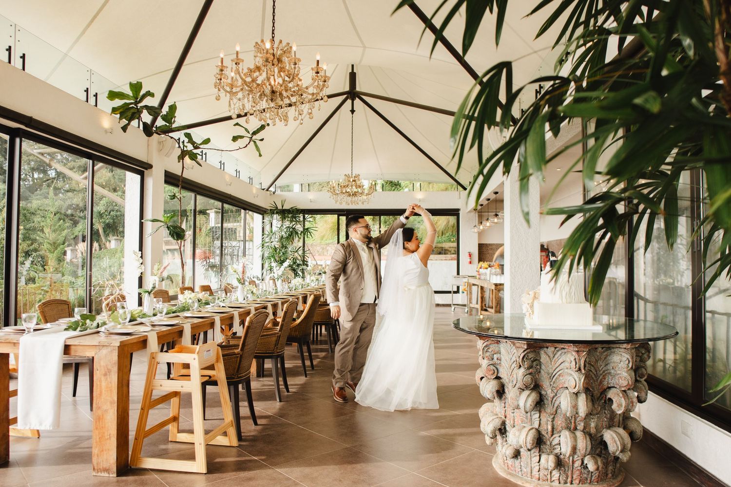 Elegant wedding venue interior with crystal chandeliers, wooden tables and chairs, and large windows overlooking greenery.