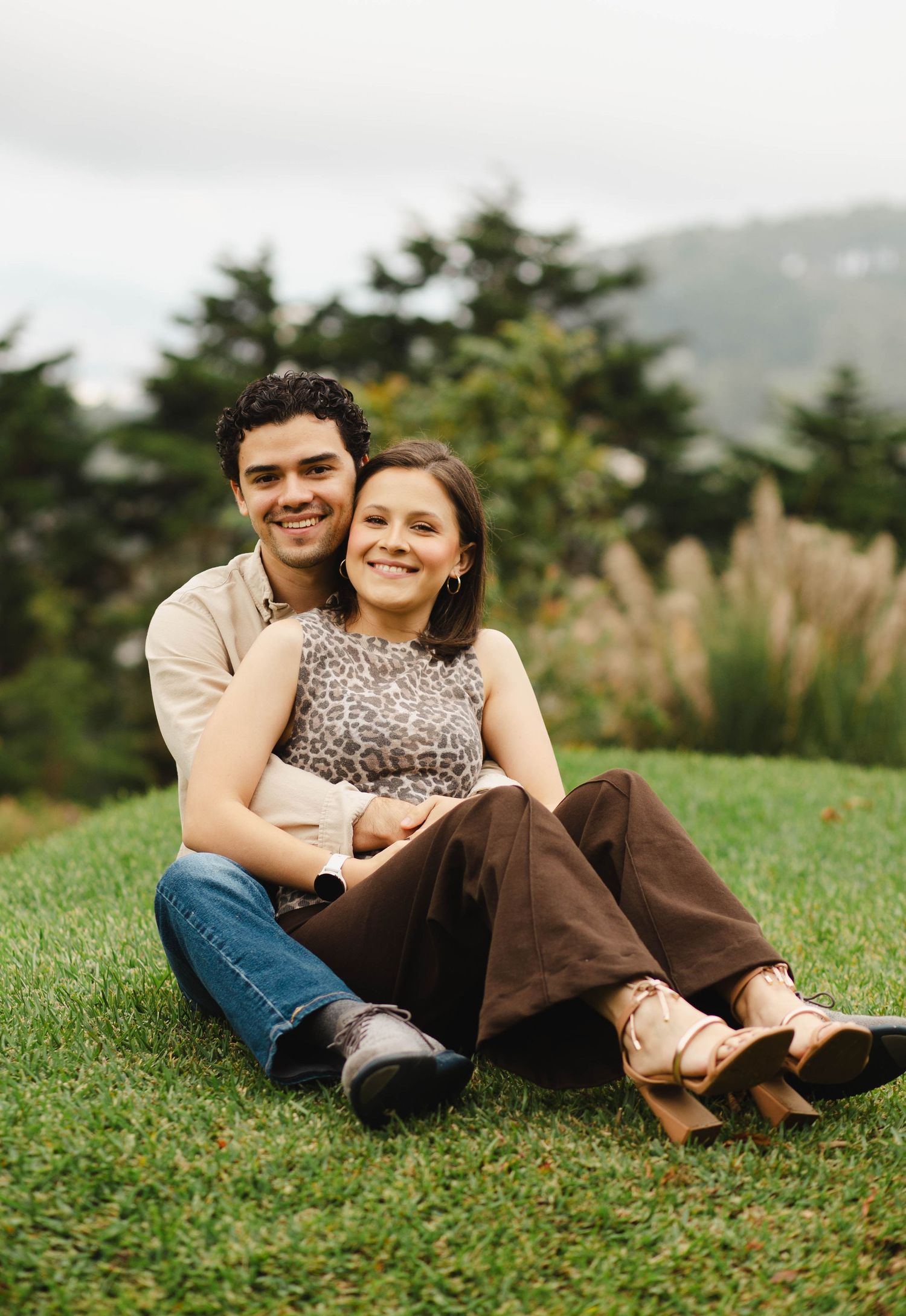 A couple sits together on green grass with evergreen trees and mountains visible in the background on a cloudy day.