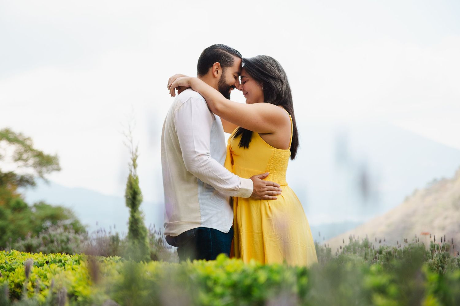 Couple in a romantic embrace outdoors, woman in yellow dress and man in white shirt, surrounded by lush greenery.