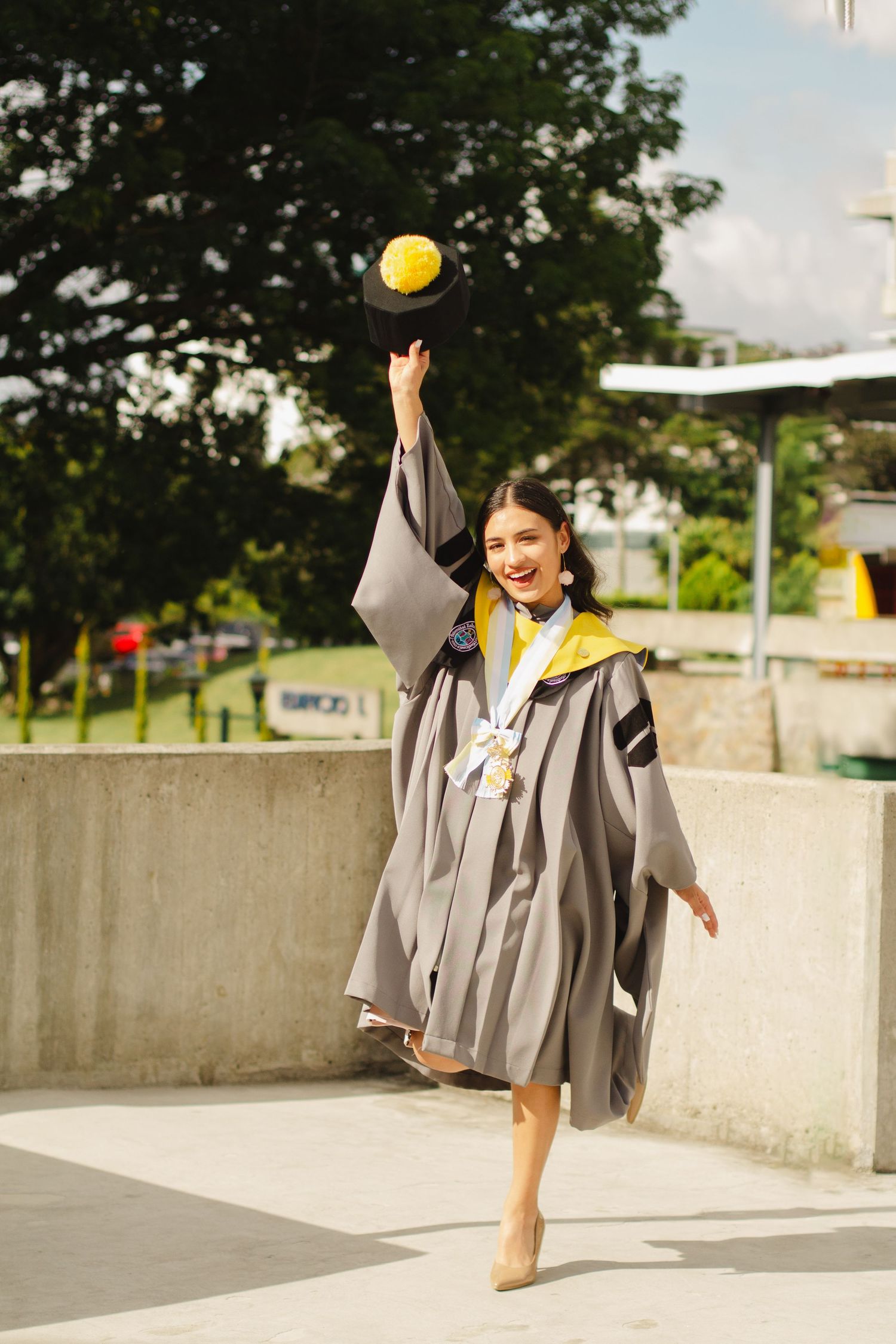 Graduate in grey academic gown tosses yellow ball in celebratory outdoor pose.