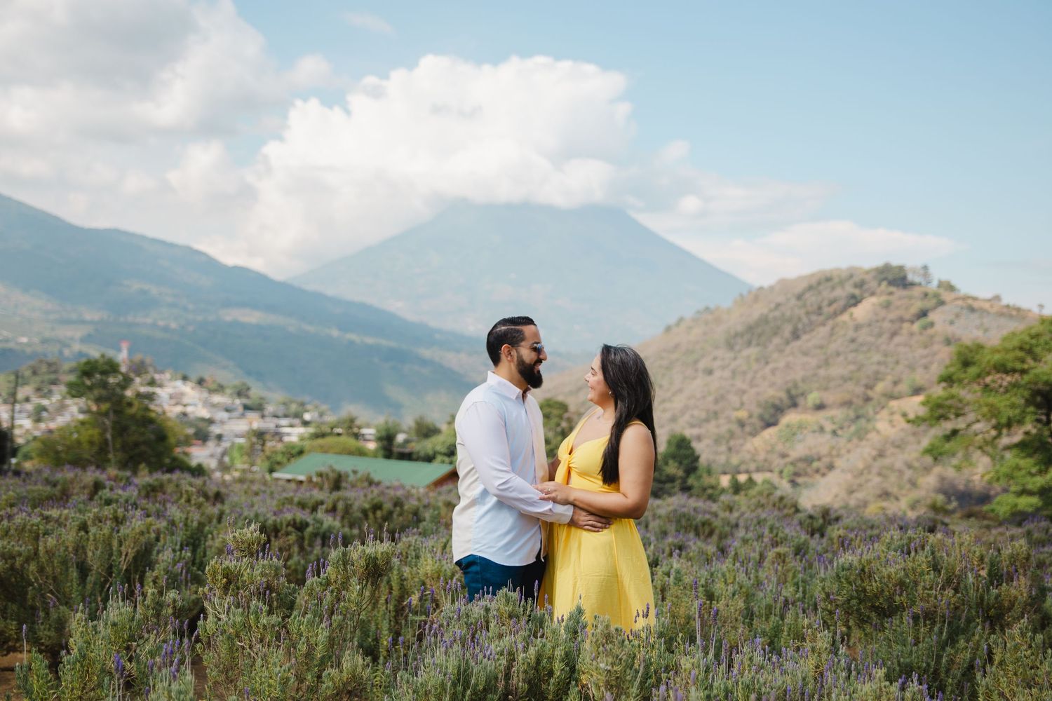 Couple in yellow and white holding hands in a field with mountains and blue sky in the background.