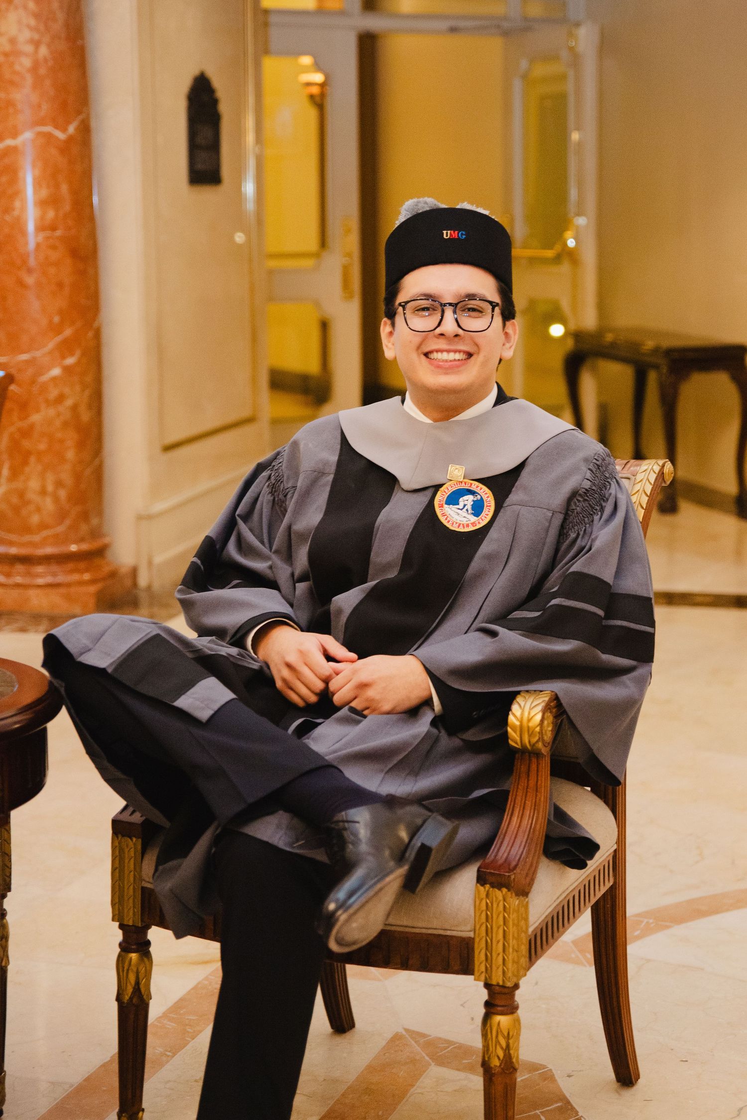 Smiling graduate in gray doctoral regalia and cap sits in ornate gold chair at formal graduation ceremony.