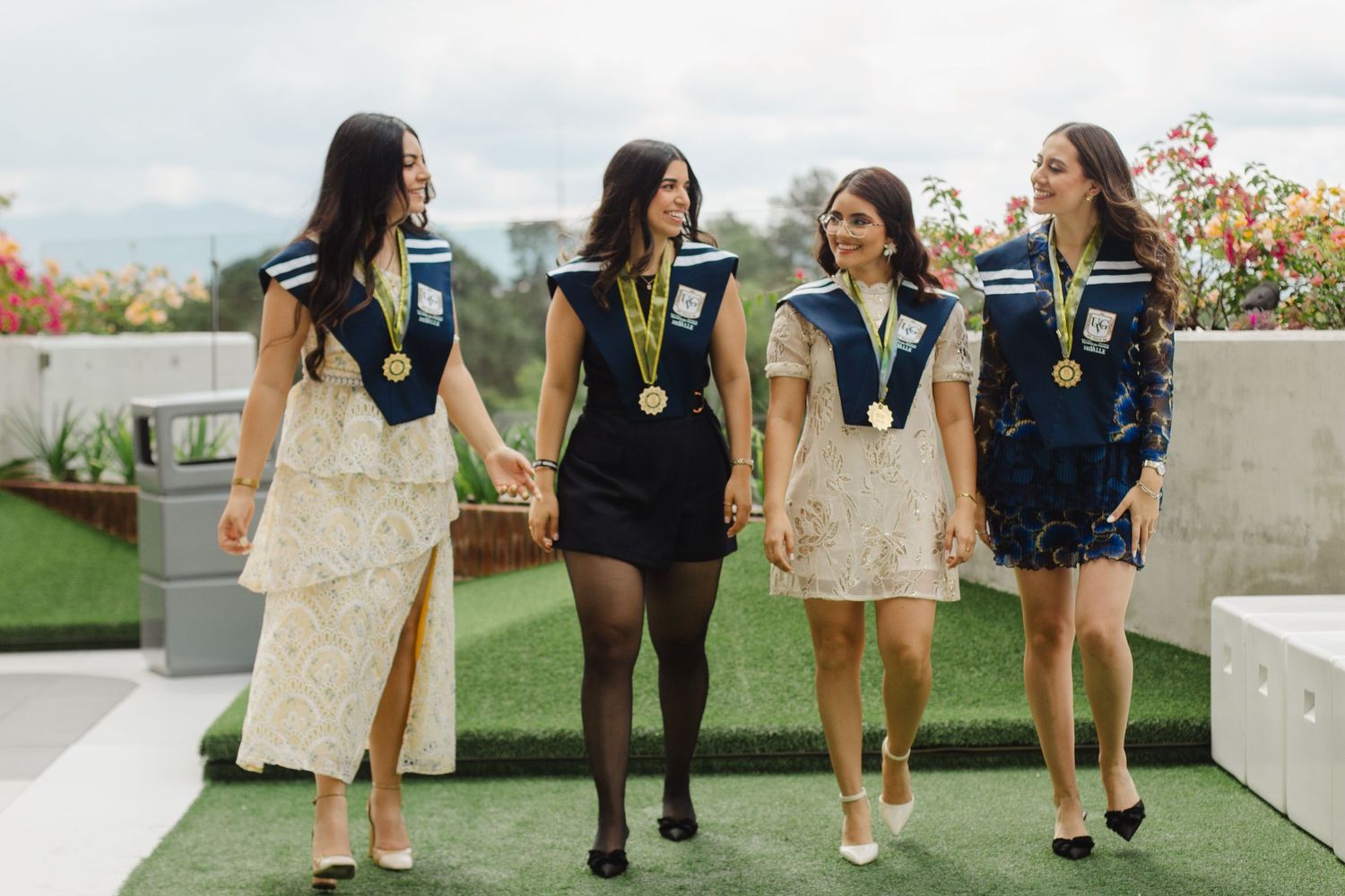 Four students wearing graduation medals and black sashes walk together on a rooftop garden space in celebratory mood.