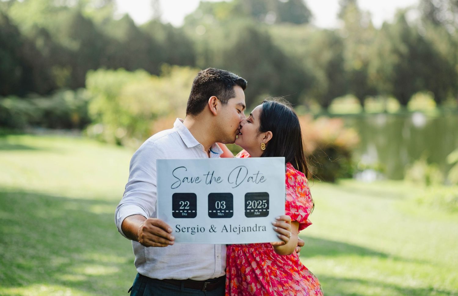 A couple holding a Save the Date sign shares an intimate moment in a sunny garden setting.