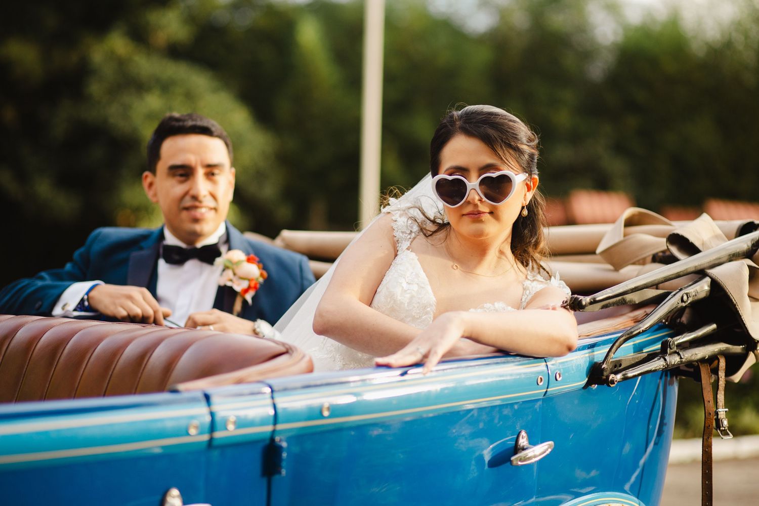 Newlyweds sitting in classic blue convertible car wearing sunglasses.
