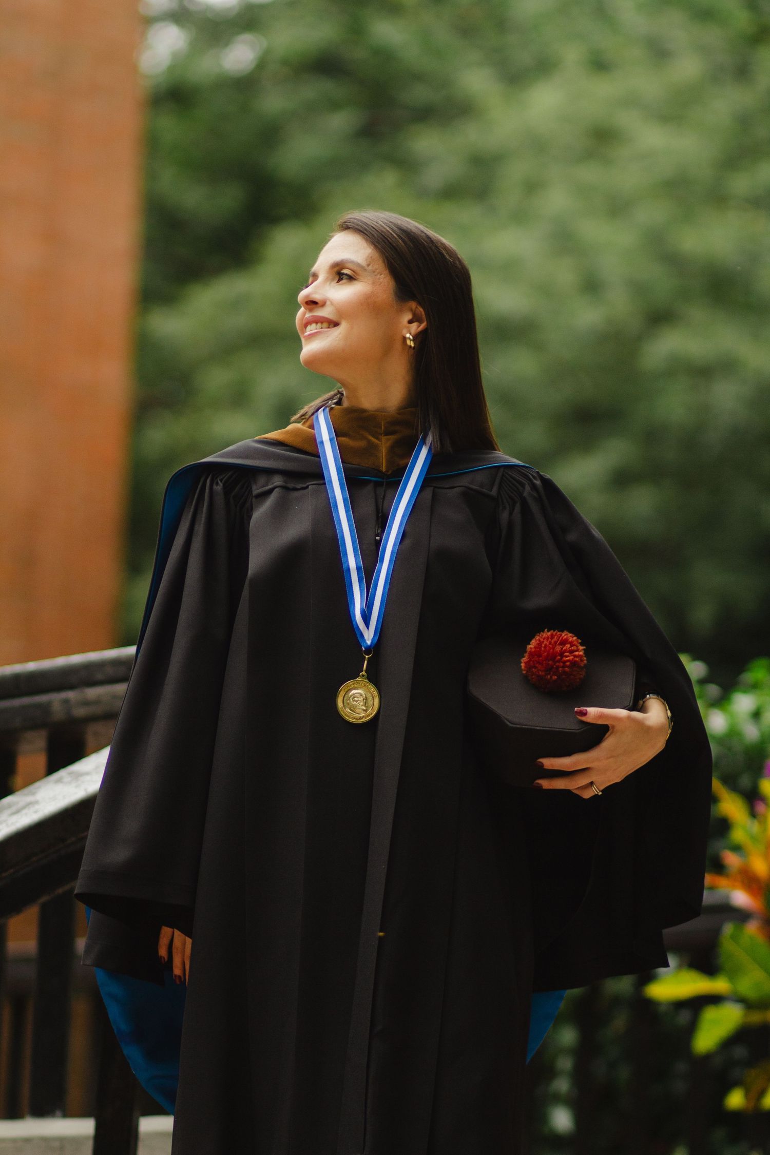 Graduate in black commencement gown stands proudly outdoors holding graduation cap with red tassel.