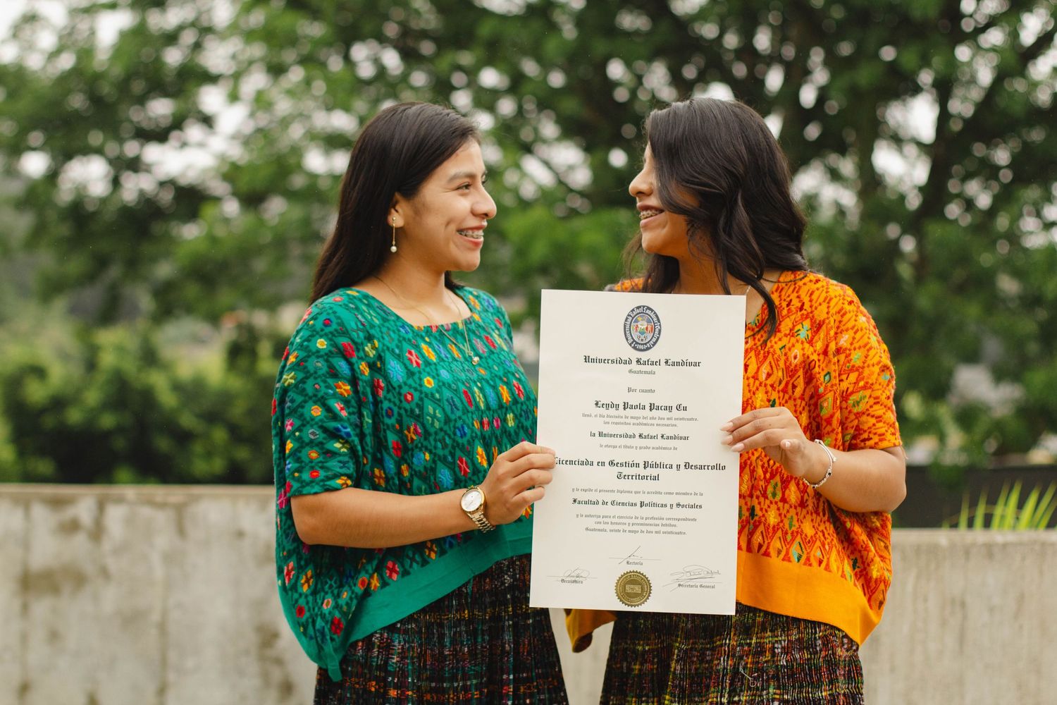 Two graduates in colorful traditional dress celebrate with diplomas outdoors.