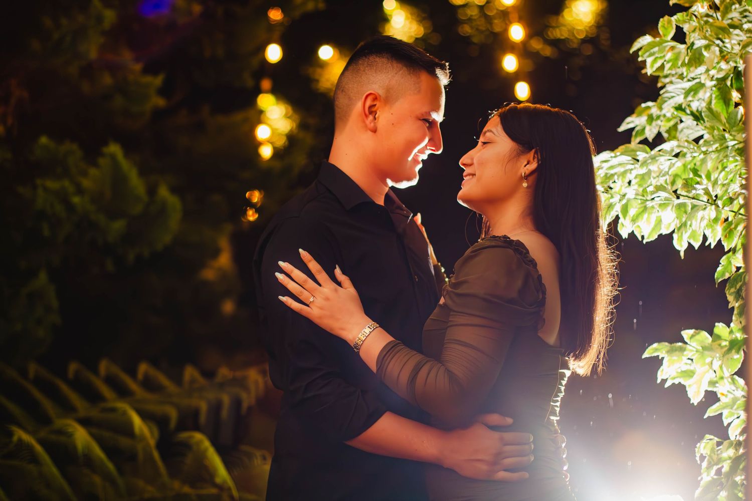 Romantic nighttime portrait of couple embracing under twinkling lights and foliage.