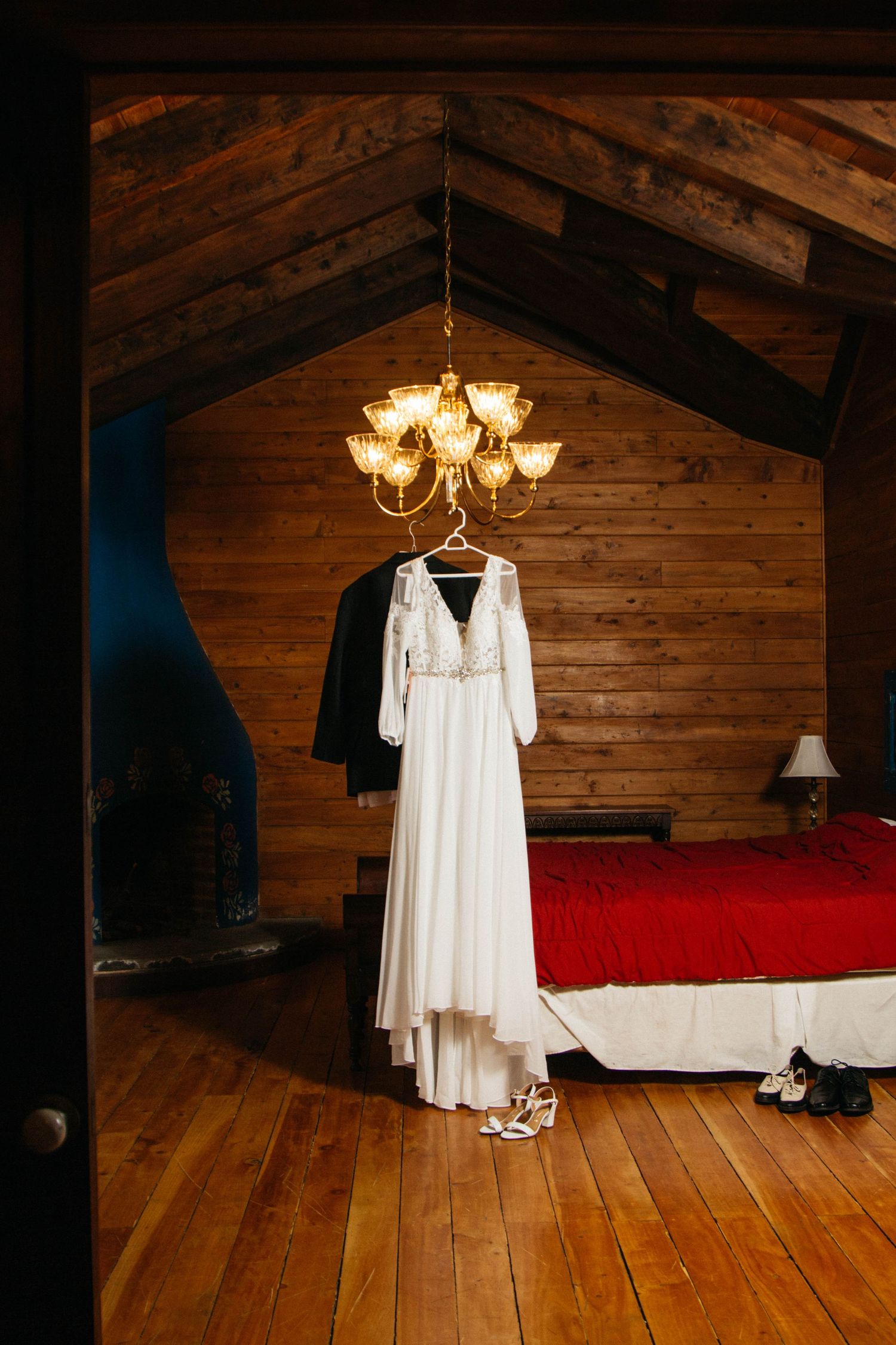 White wedding dress hanging beneath chandelier in rustic wooden cabin bedroom.