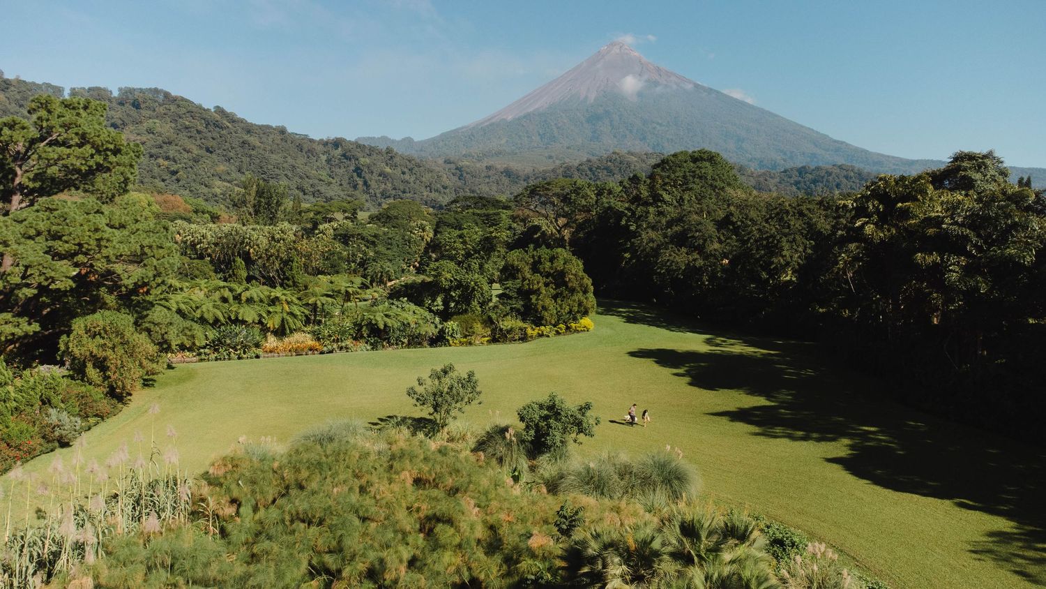 A panoramic view of a volcano rising above green fields and forested hills under blue skies.