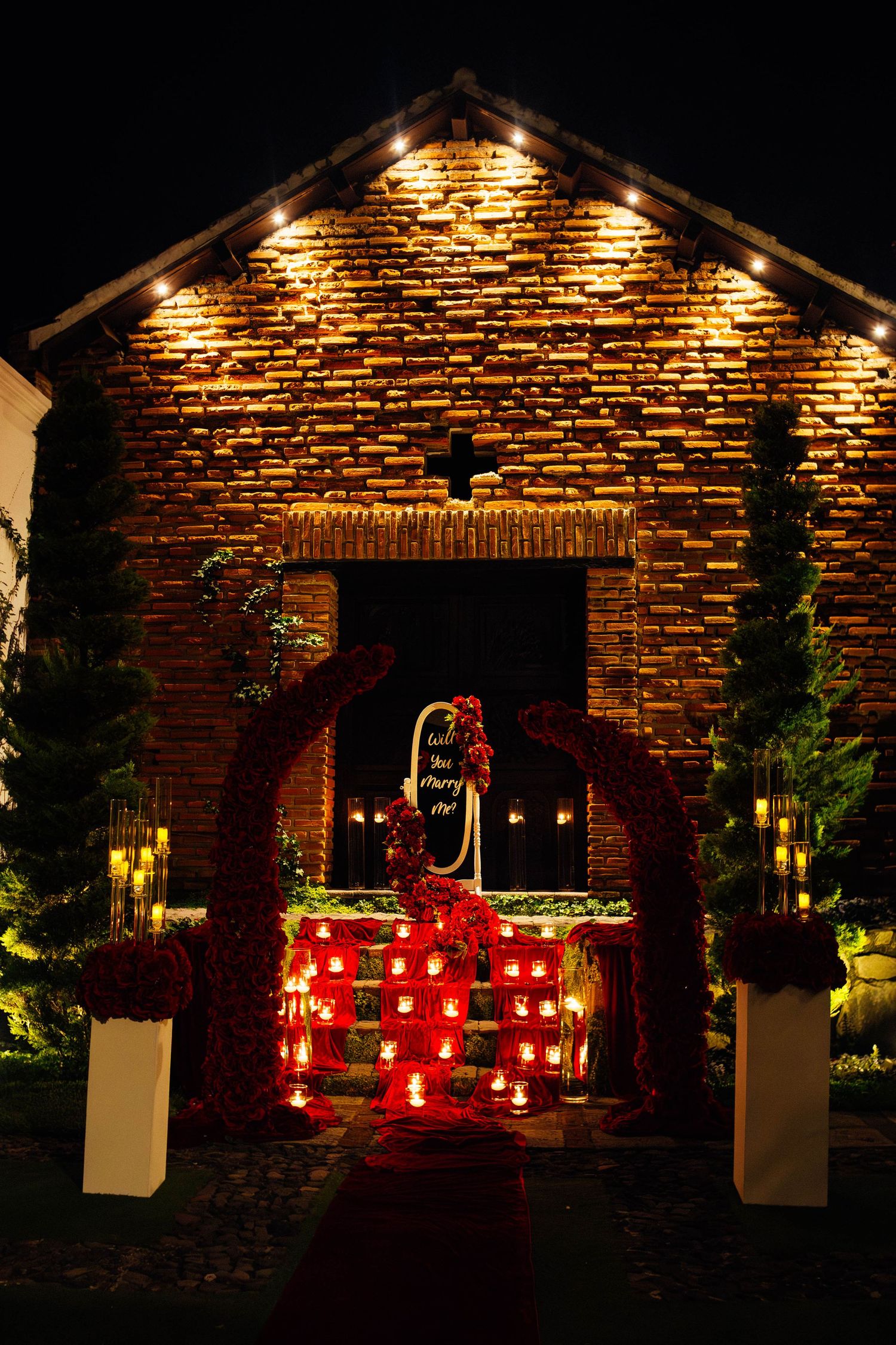 Illuminated brick chapel entrance decorated with red candles and string lights for romantic proposal.