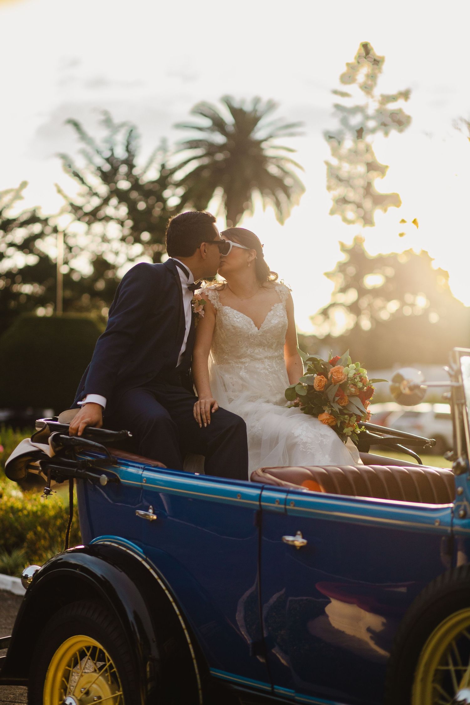 A romantic sunset wedding photo featuring a couple seated in a vintage blue convertible car with palm trees in the background.