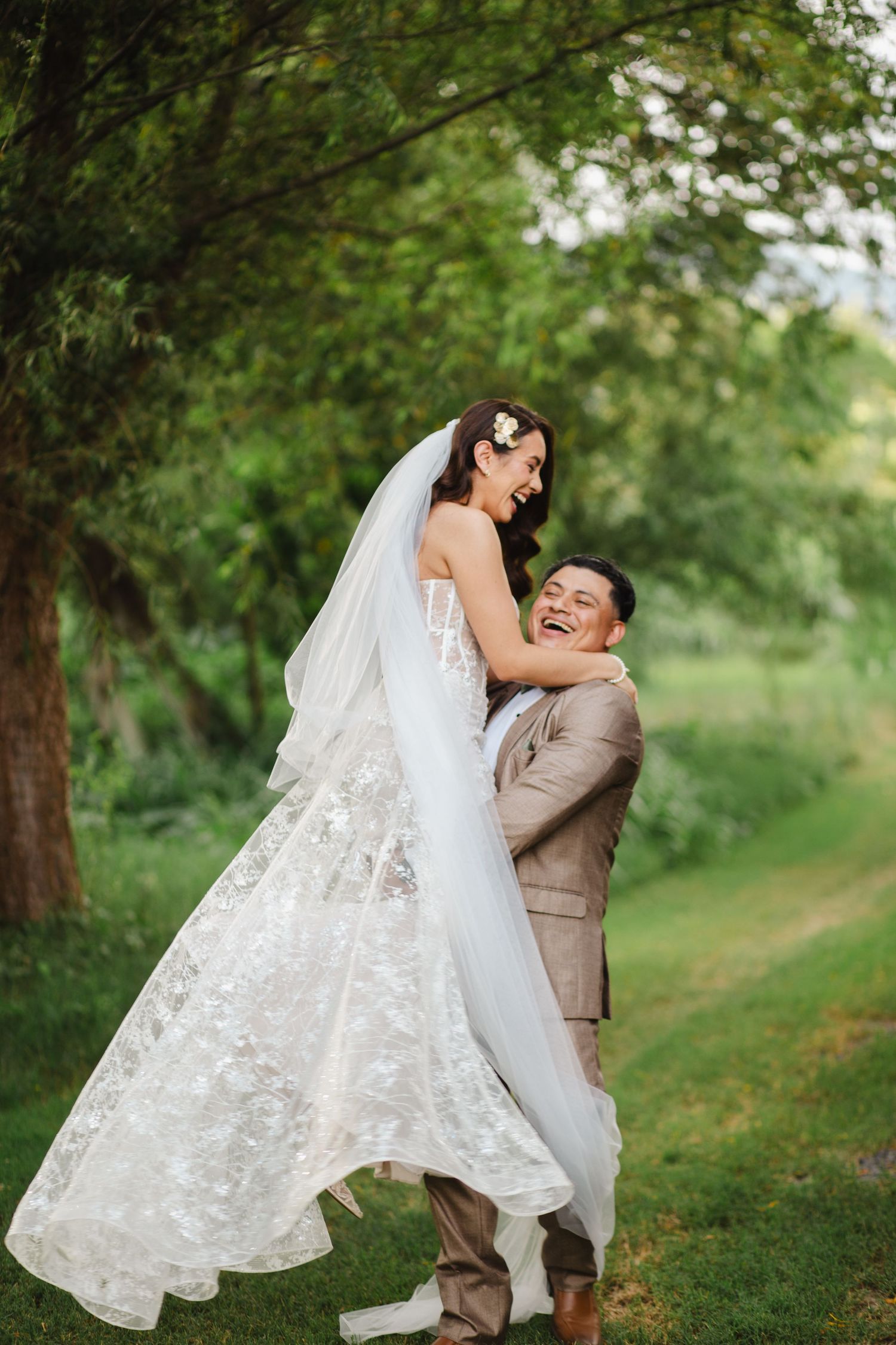 Joyful groom lifting smiling bride in lace wedding dress and veil outdoors among lush green trees.