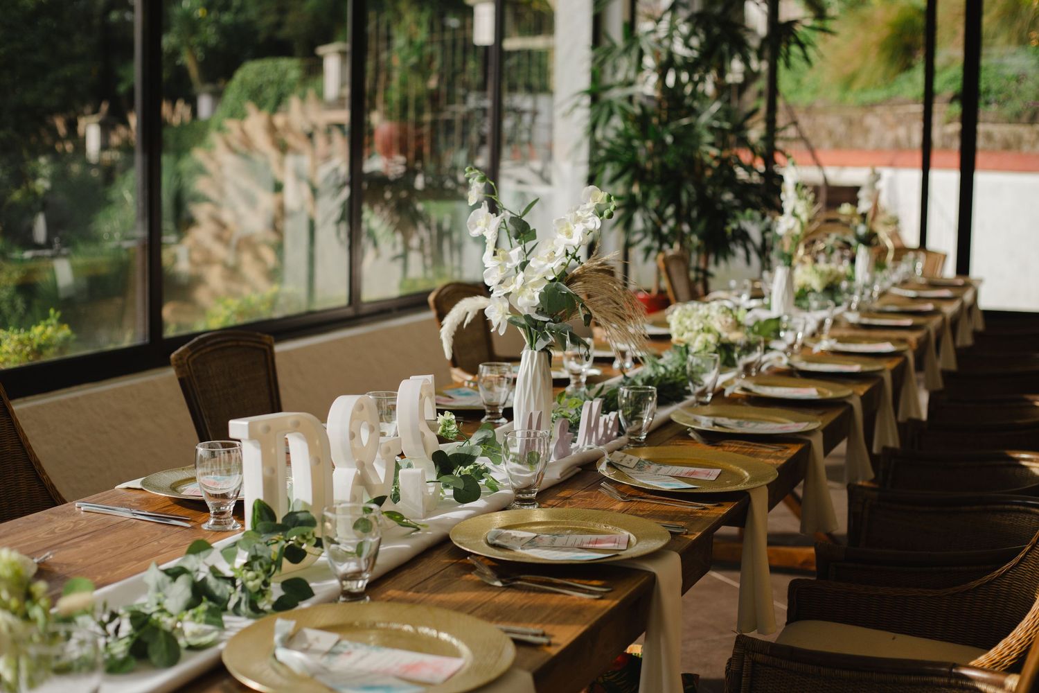 Long wooden dining table decorated with elegant place settings and greenery in a rustic restaurant setting.