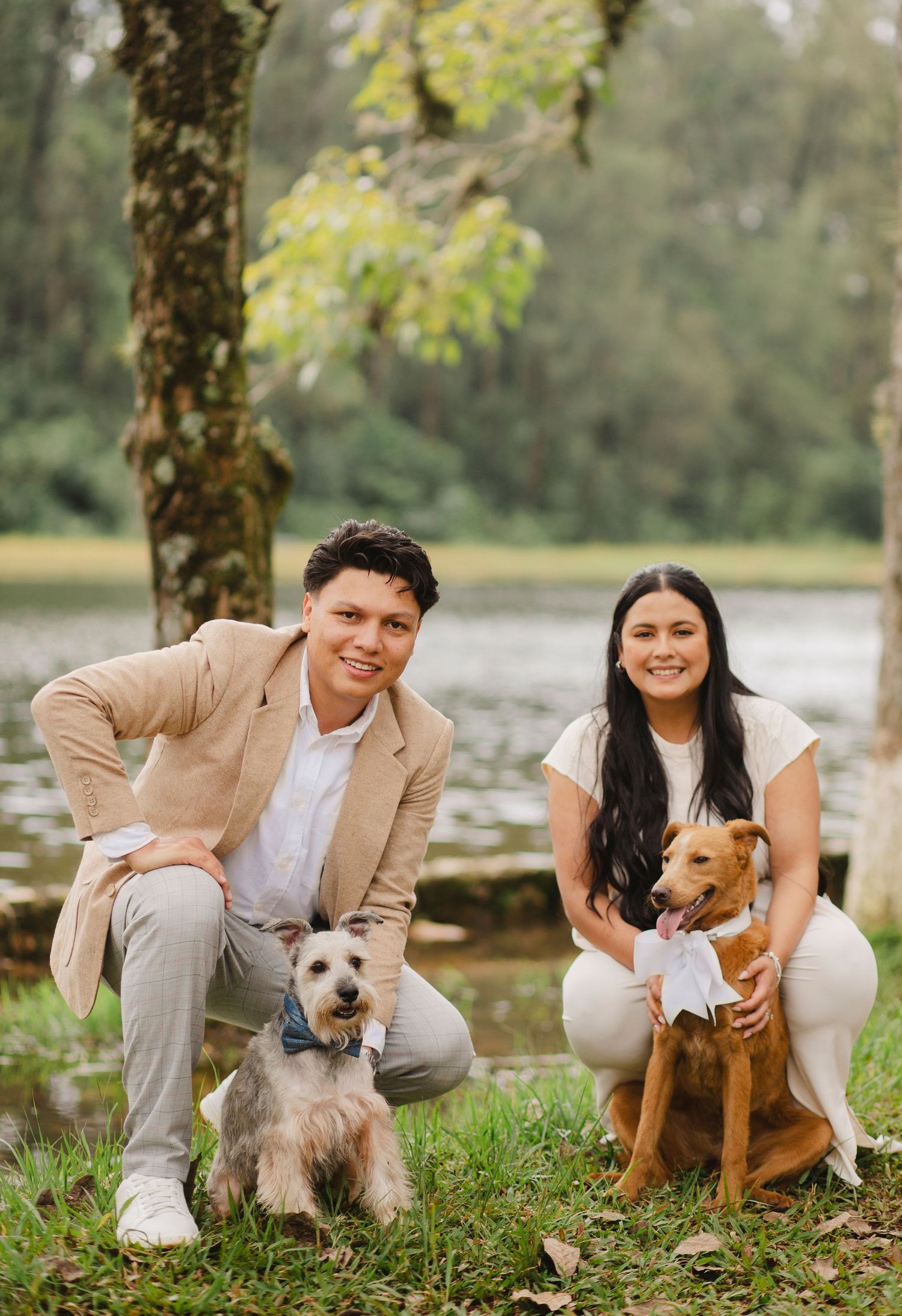 A couple sits outdoors with their two dogs on grass near a lake during a photo session.