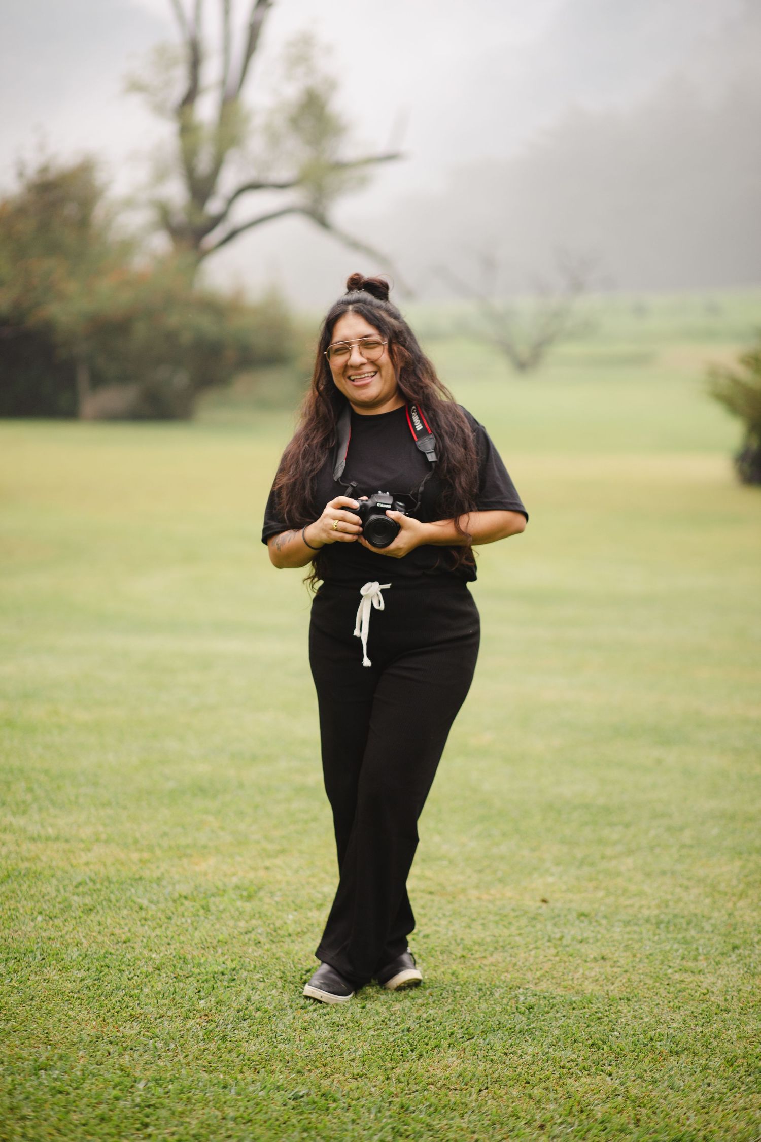 A photographer in black clothing captures photos while posing on a misty green lawn.