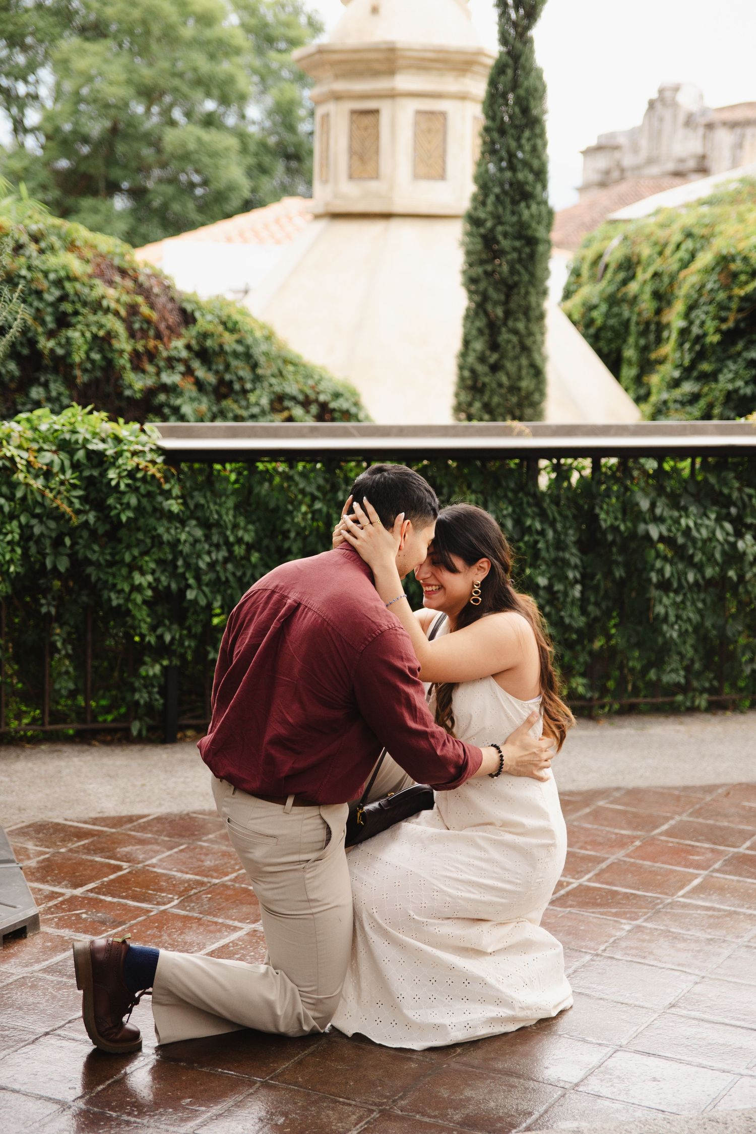 Couple sharing an emotional embrace on one knee during a marriage proposal on a terrace surrounded by lush greenery.