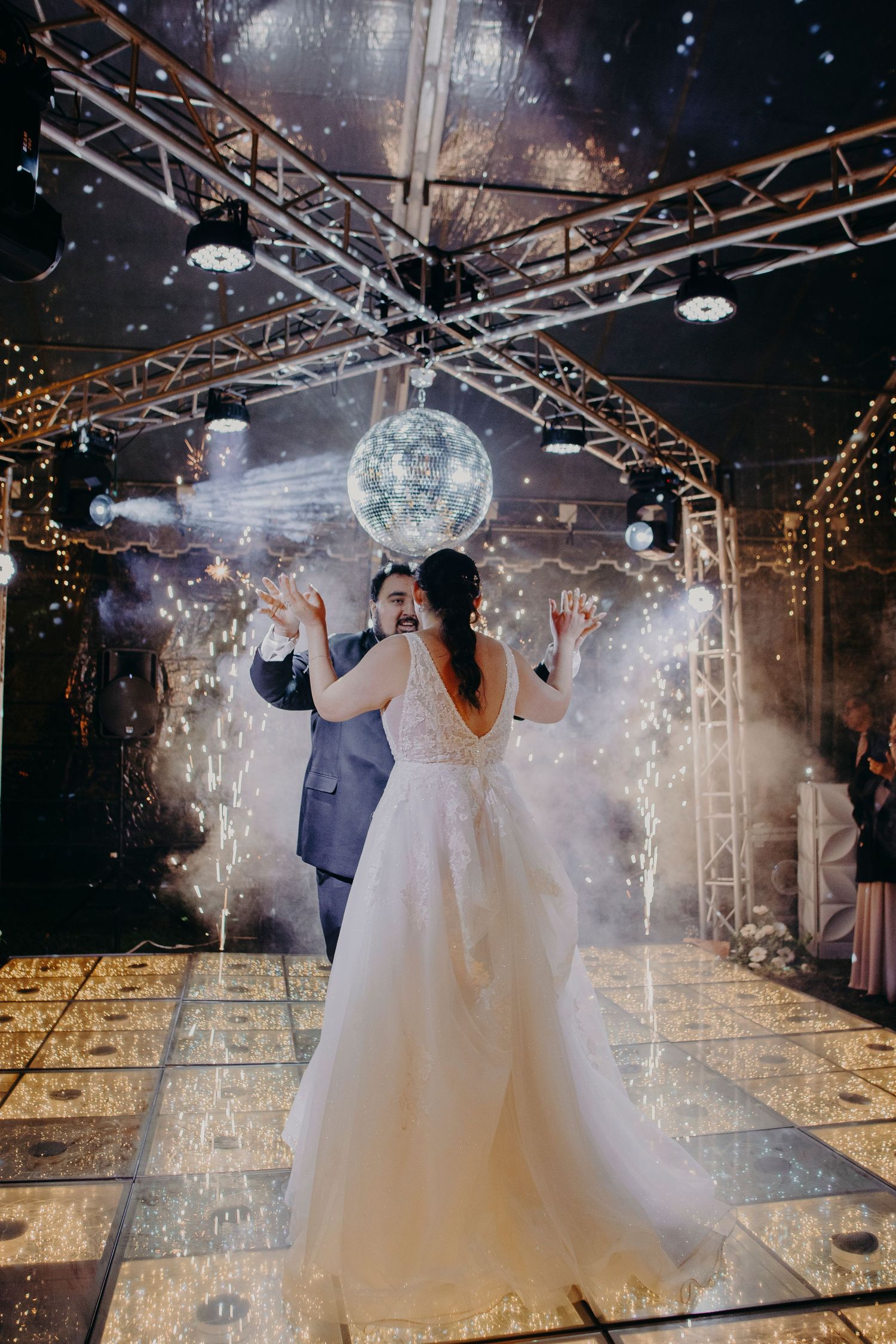Couple dancing on a reflective floor under twinkling lights and disco ball in an elegant wedding venue.