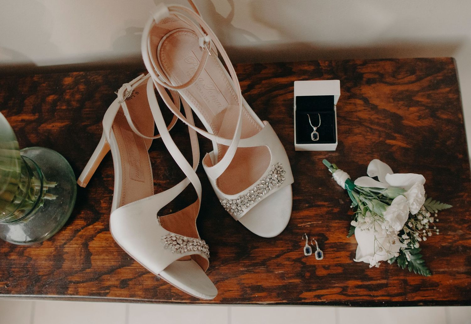 Wedding accessories including sparkly white heels, engagement ring box, and delicate floral boutonniere on rustic wooden surface.