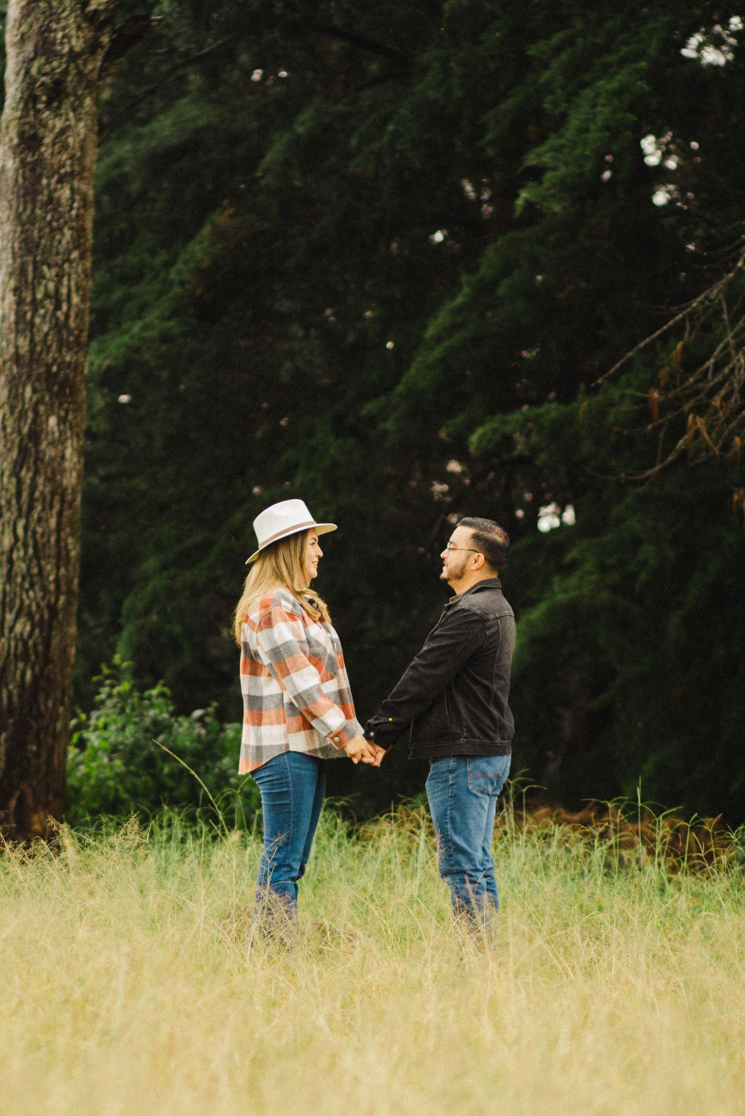 Couple holding hands while standing face to face in golden field with forest backdrop.