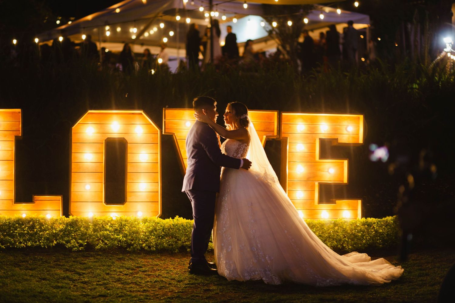 A couple embraces in front of illuminated LOVE marquee letters at a nighttime outdoor wedding reception.